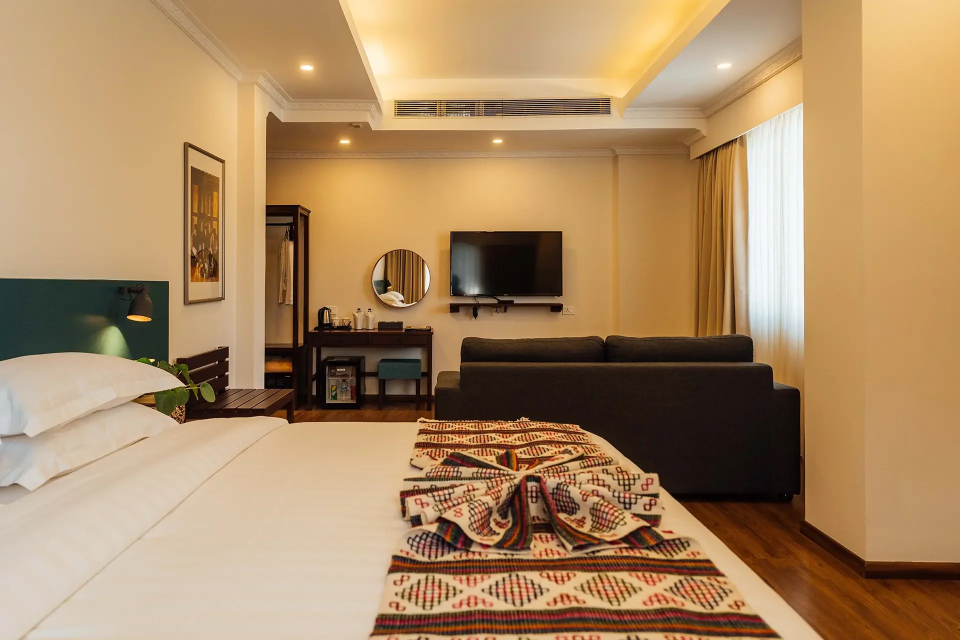 Hotel room with bed, black sofa, wall-mounted TV, mirror, art print, window with curtains, and a small wooden table.