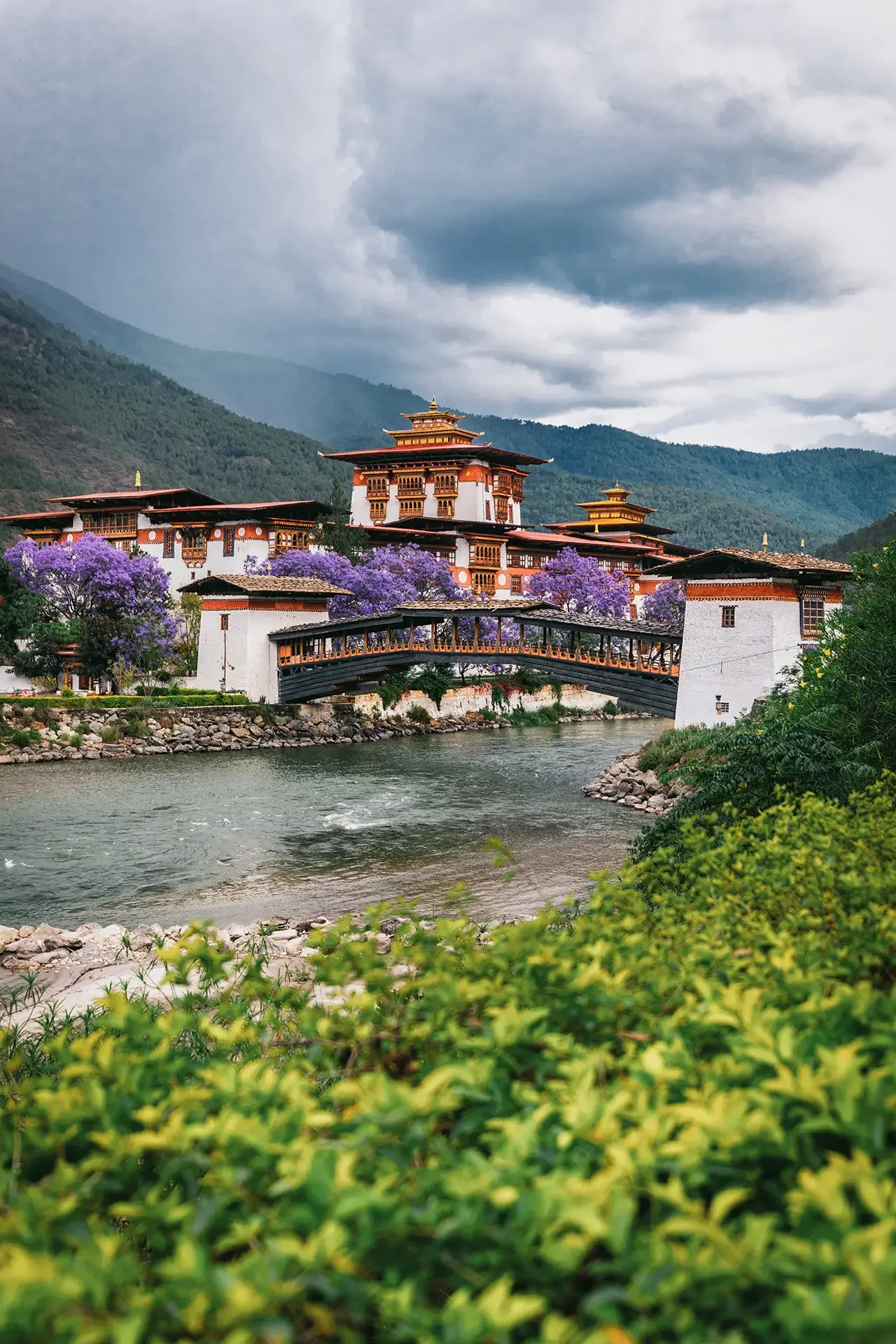 View of a traditional Asian-style building complex with multiple tiered roofs, situated by a river with a stone bank. The scene includes lush green trees, purple flowering trees, and a backdrop of forested mountains under a cloudy sky.