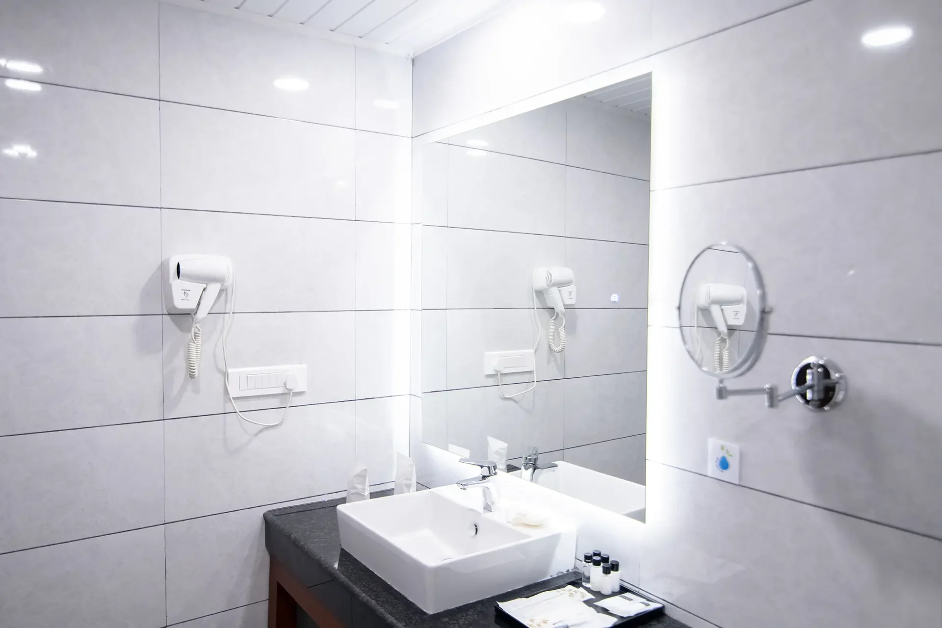 A modern bathroom sink area with a large mirror, white tiled walls, and a black countertop. There are two white hairdryers mounted on the wall and a round magnifying mirror attached to the wall. The sink has a faucet and three bottles of toiletries on the countertop.