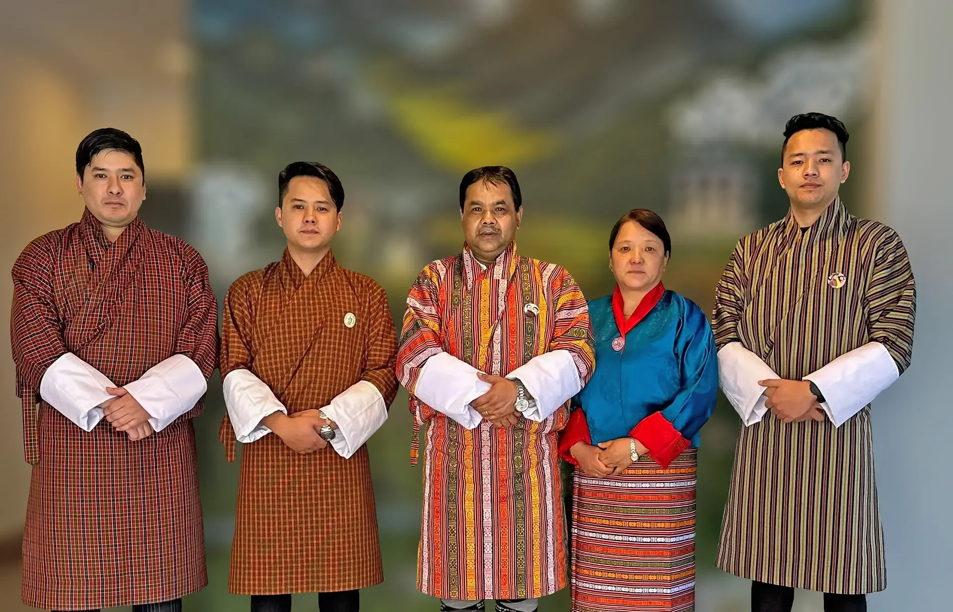 A group of five people standing side by side, dressed in traditional Bhutanese attire called gho for men and kira for women, posed against a blurred outdoor background.