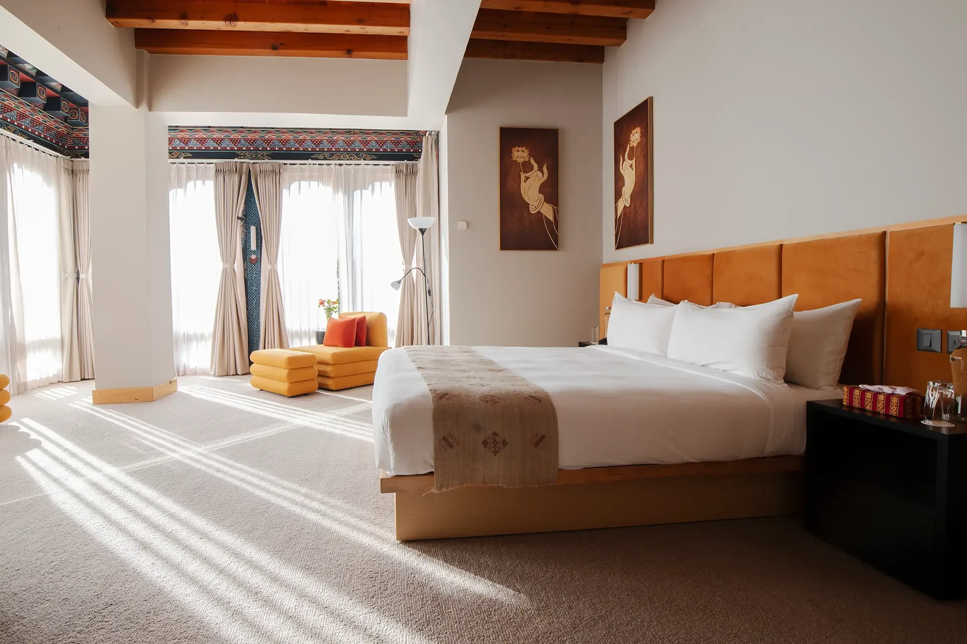 A hotel room with a large bed, wooden headboard, beige walls, and windows with cream curtains. There is a yellow armchair with orange cushions, a white floor lamp, and framed artwork of lotus flowers on the walls.