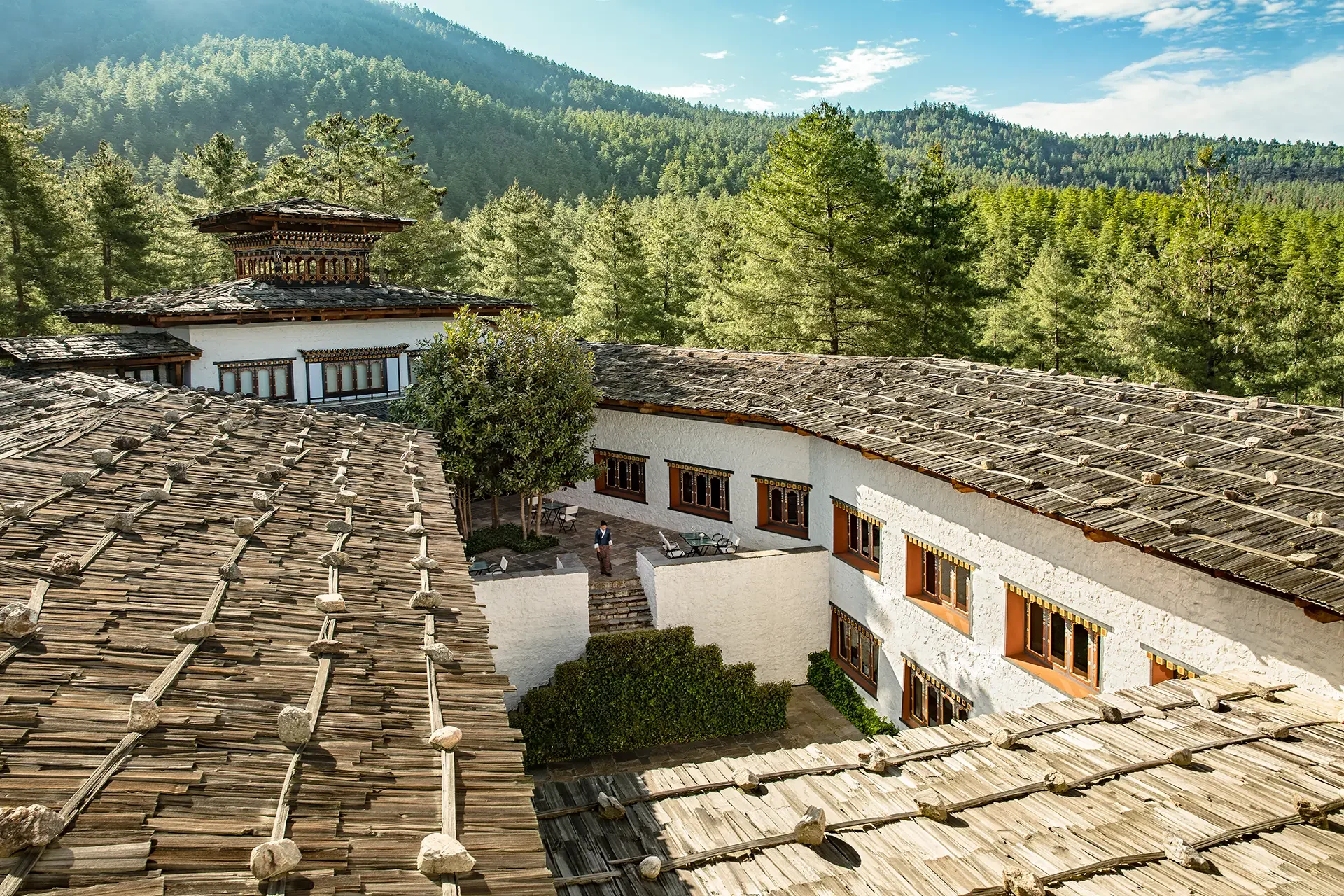 View of traditional Bhutanese buildings with wooden tile roofs beneath forested hills and blue sky.