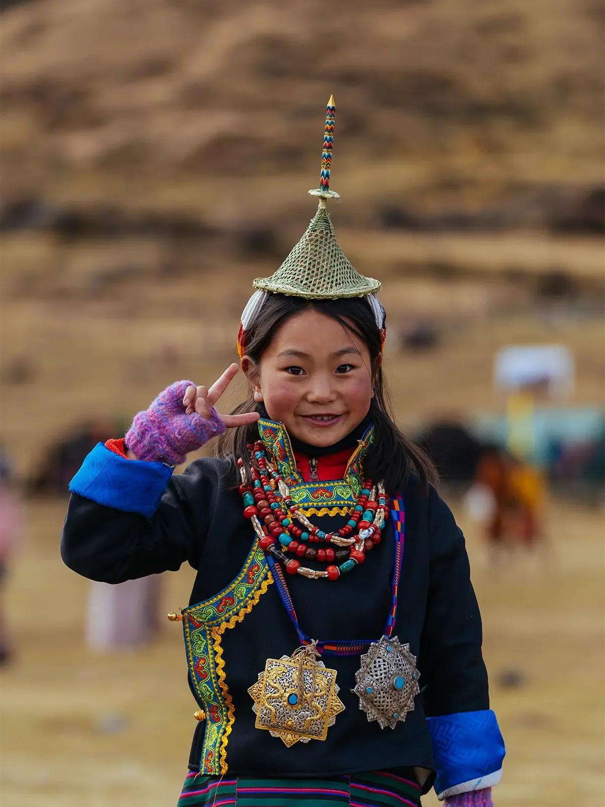 A young girl in traditional clothing and jewelry, making a peace sign and wearing a hat with a decorated pencil on top, smiling in an outdoor setting.