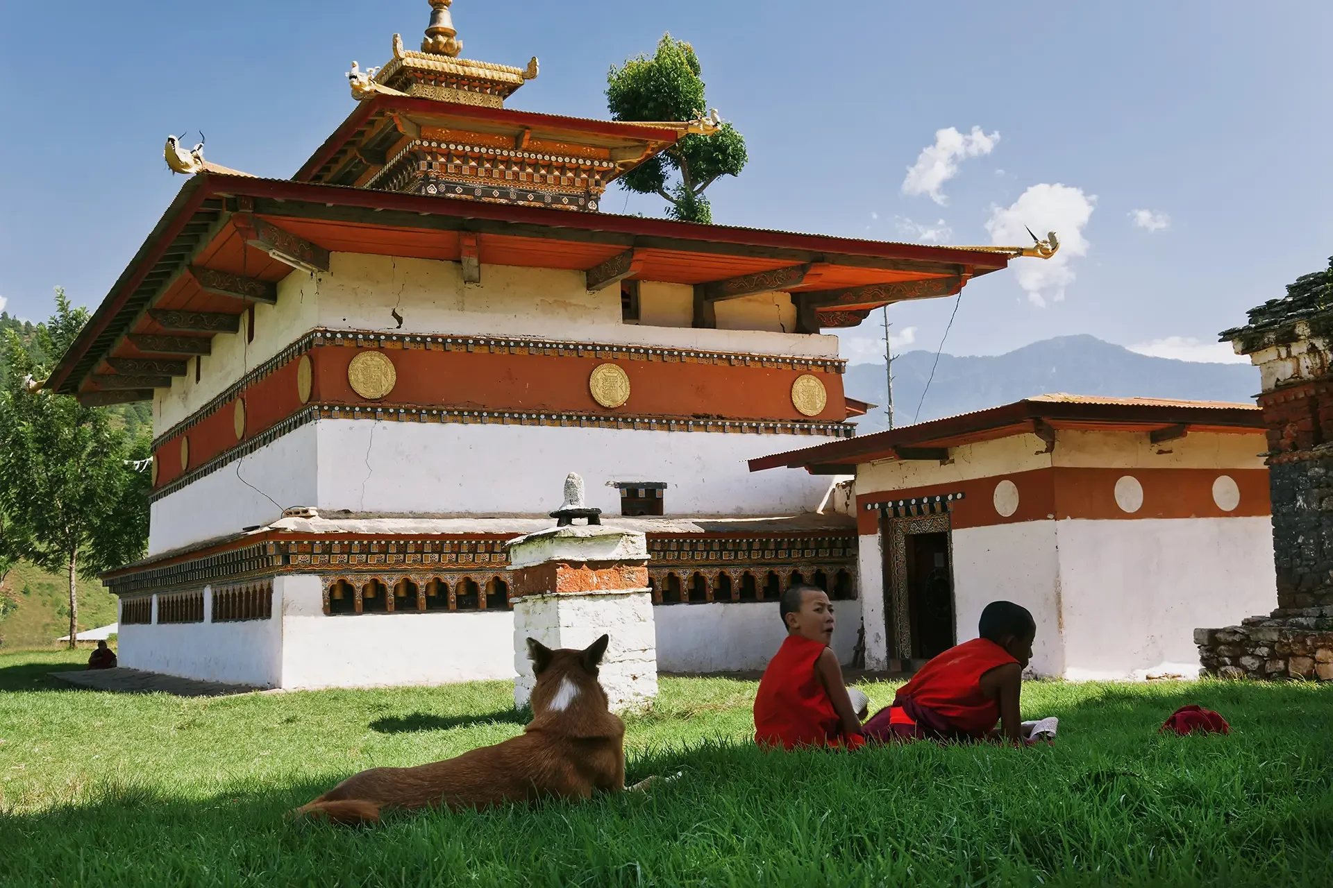 A traditional Tibetan temple with red, white, and gold decorations, situated in a green landscape with mountains in the background. Two children in red clothing sit on the grass nearby, with a dog lying in the foreground.