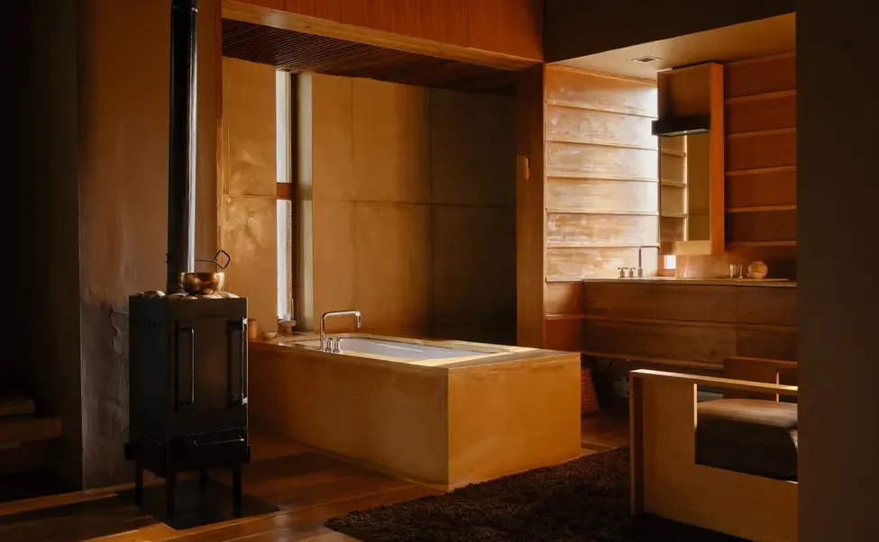 Interior of a cozy, wood-paneled room with a built-in bathtub, a small wood stove, and minimal decor, featuring warm lighting and a rustic aesthetic.