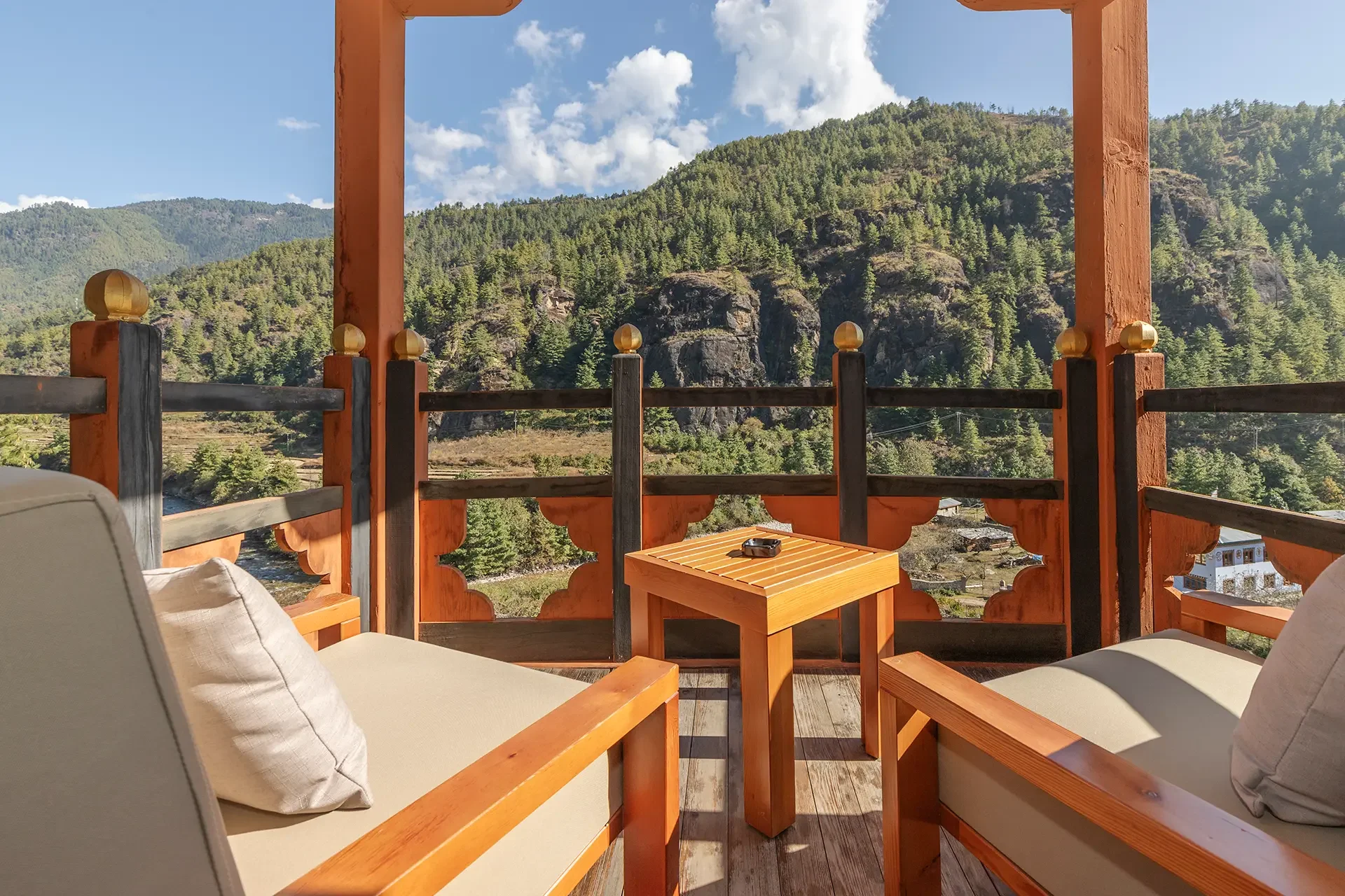 View from a balcony with two cushioned chairs and a wooden table, overlooking a scenic mountainous landscape with green trees, rocky cliffs, and a partly cloudy sky.
