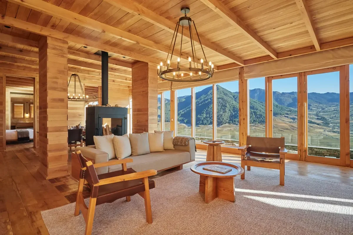 Living room with wooden walls, ceiling, and floor, featuring large windows with mountain views, a beige sofa, two wooden chairs, a small wooden side table, and a round coffee table with magazines, illuminated by a circular chandelier.