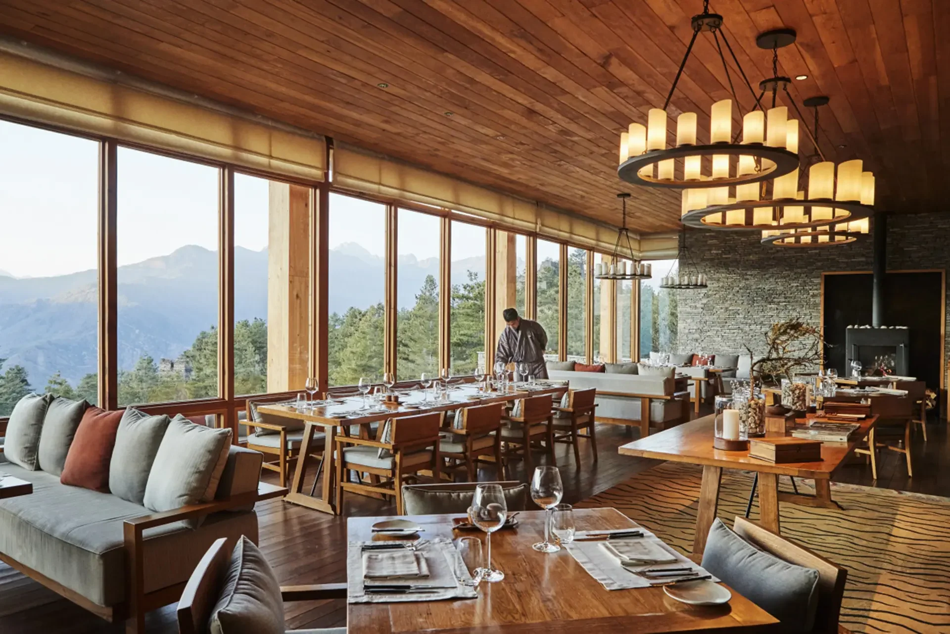 Elegant dining area with large windows overlooking mountains, wooden ceiling with modern chandeliers, and a mix of seating including a sofa with cushions and tables set for a meal.