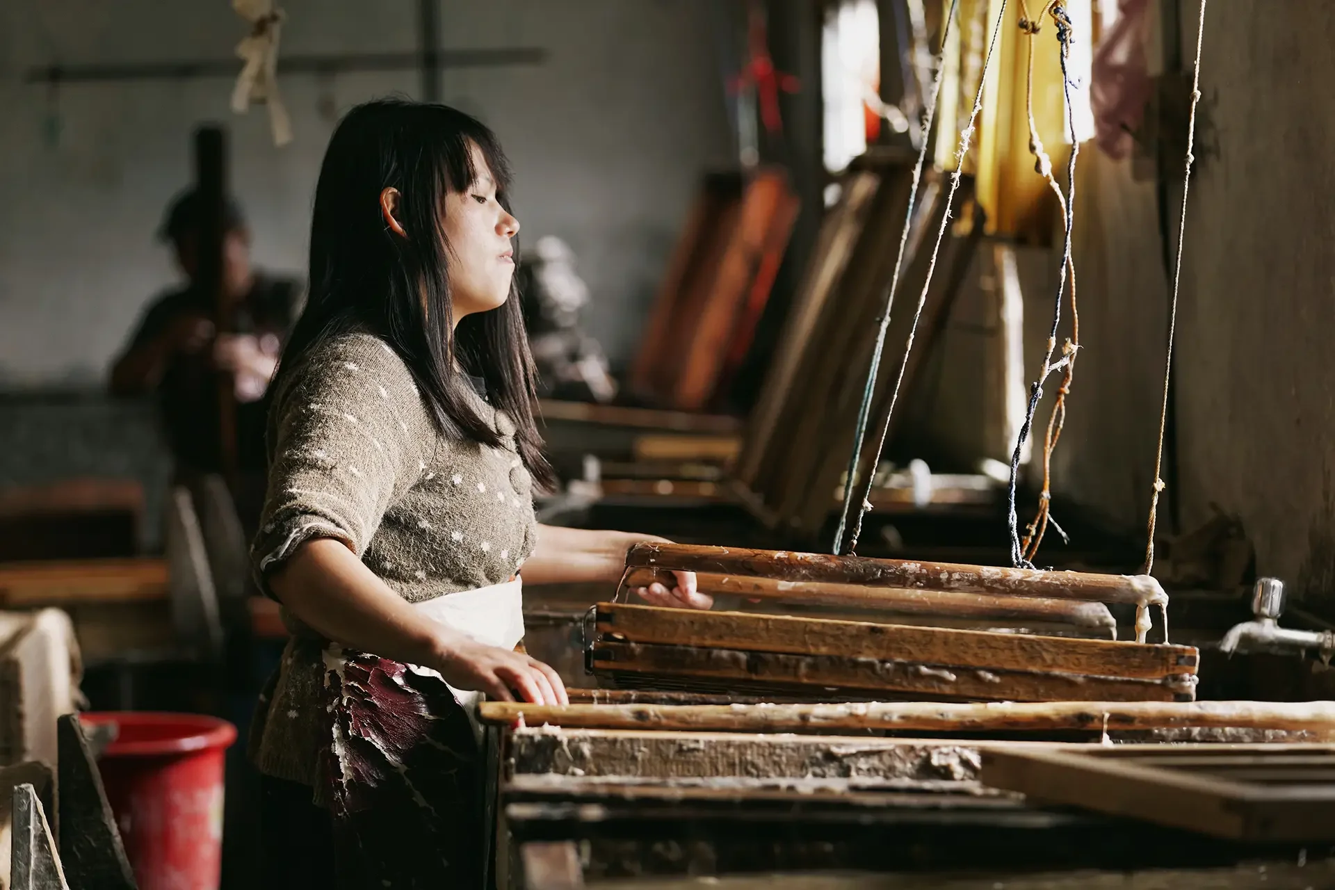 A woman with black hair in a brown sweater works at a wooden loom in a workshop with natural light coming through a window.