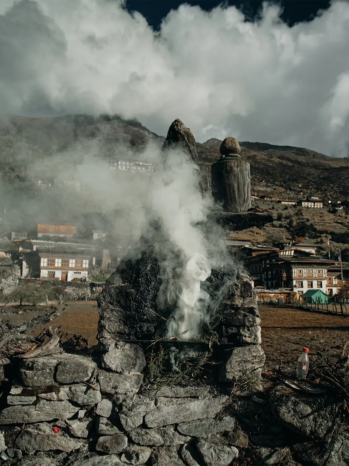 A stone fire pit with smoking incense sticks surrounded by stones and ruins, with mountains, clouds, and traditional buildings in the background.
