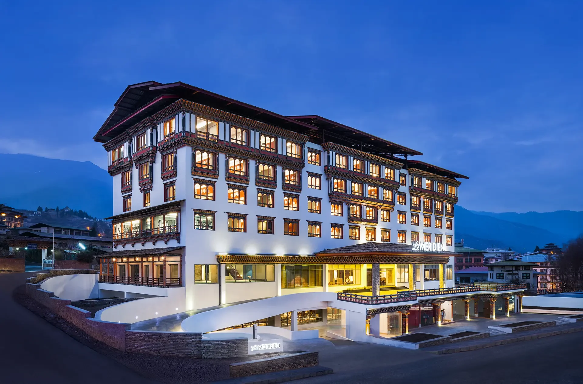 A large hotel building with multiple floors and traditional Asian architectural elements, illuminated at dusk, with mountains in the background.