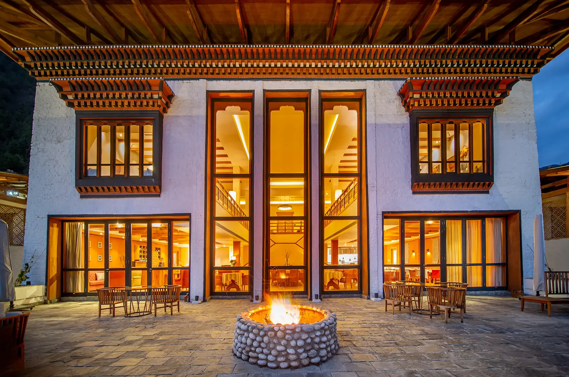 A modern two-story house with large glass windows and a white exterior, illuminated warmly from inside, with an outdoor stone-paved patio and a central fire pit in front. The house features traditional roof overhangs and intricate woodwork detail.