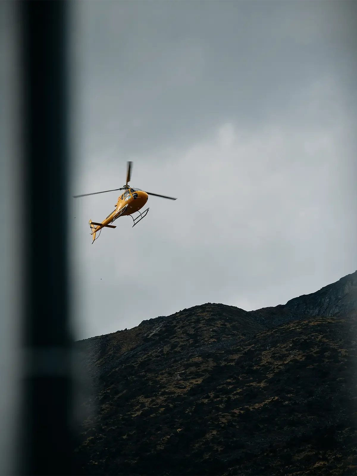Yellow helicopter flying in cloudy sky above a mountain range with dark, rugged terrain.