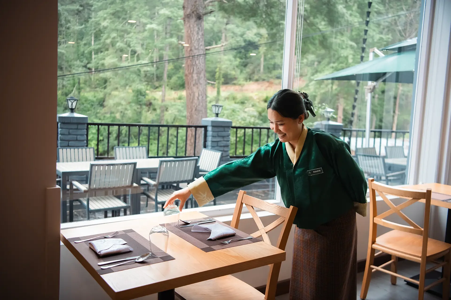 A woman wearing a dark green uniform and plaid skirt is setting a dining table inside a restaurant, with an outdoor patio and forested area visible through large windows.