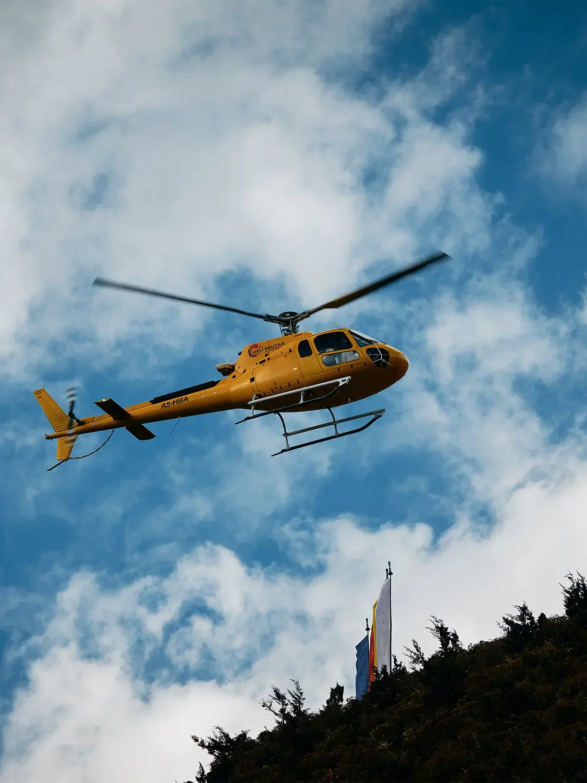 Yellow helicopter flying in a partly cloudy sky above a hillside with flags and trees.