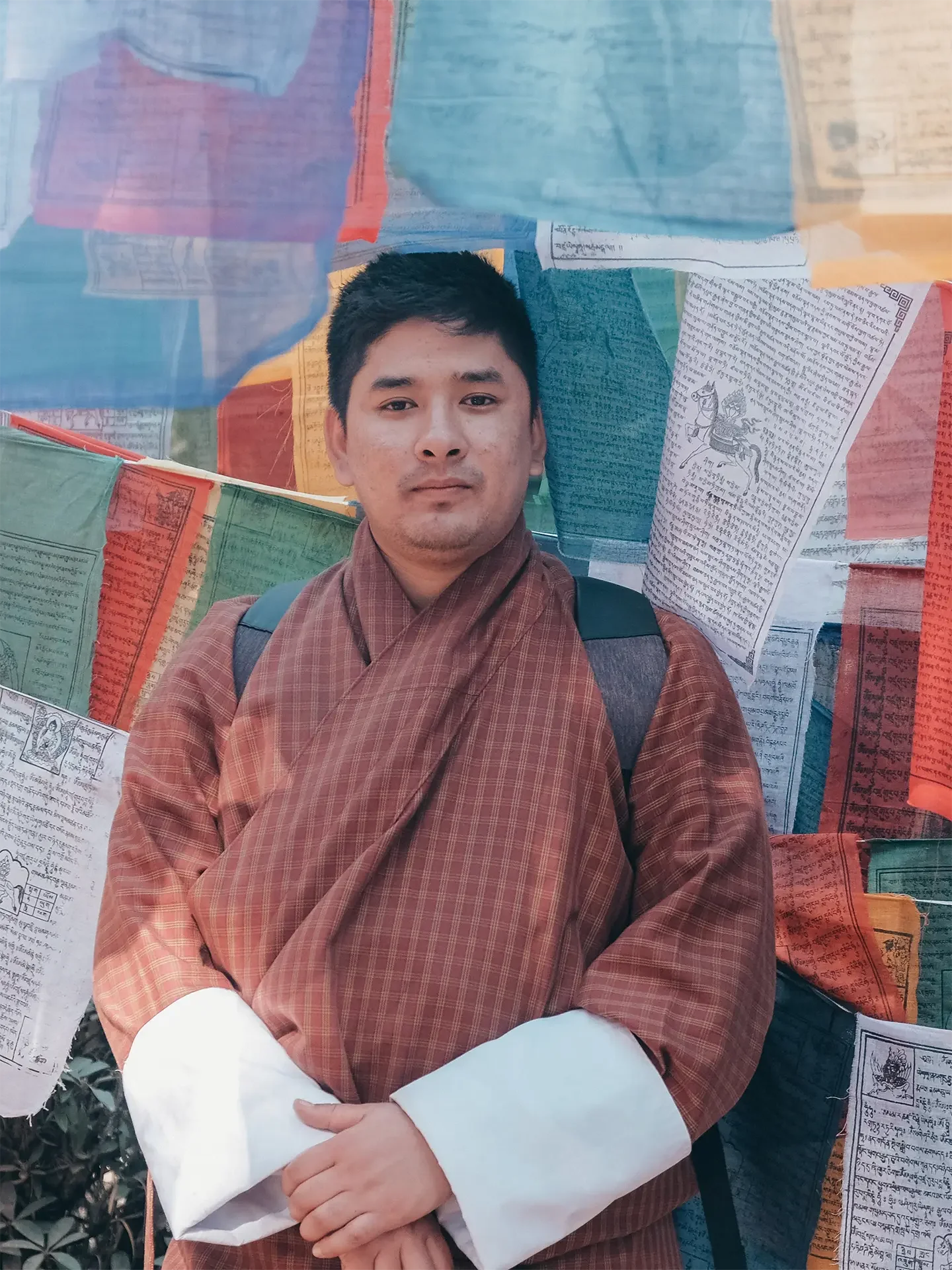 A young man in traditional orange and white clothing, standing with arms crossed in front of a backdrop of colorful prayer flags with writings and symbols.