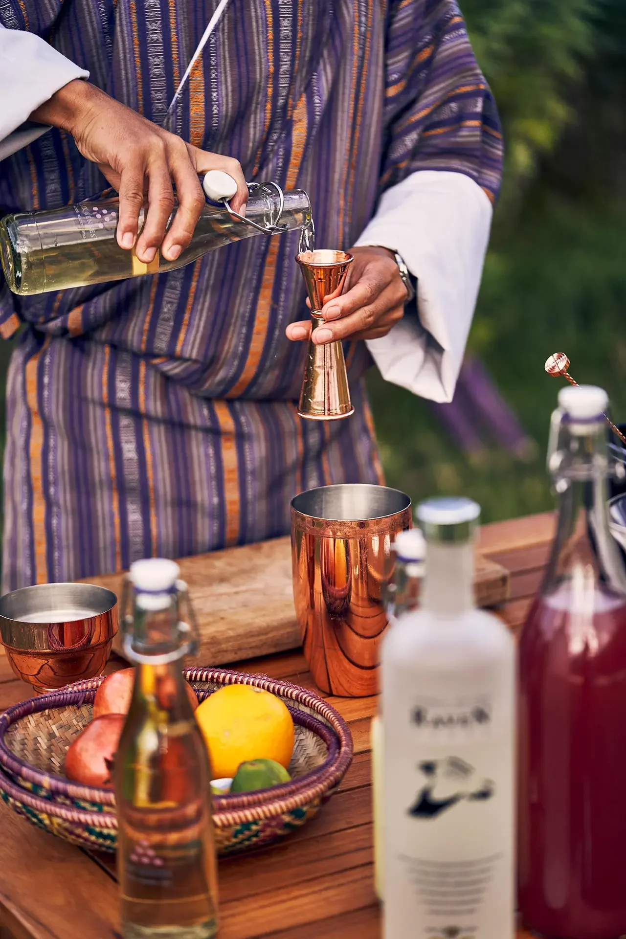Person pouring a drink into a copper jigger at an outdoor bar setup with various bottles, a basket of fruit, and other barware on a wooden table.