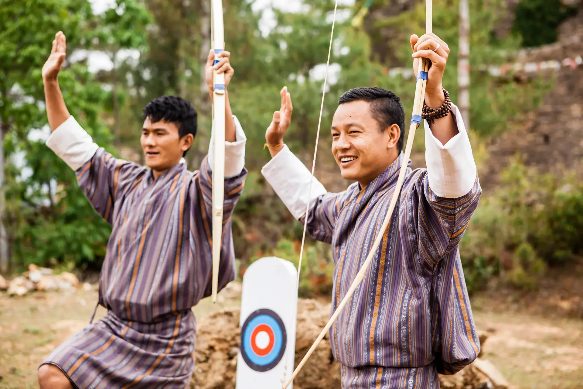 Two men in traditional Bhutanese attire celebrating outdoors beside an archery target and a white archery bow, with trees and natural surroundings in the background.