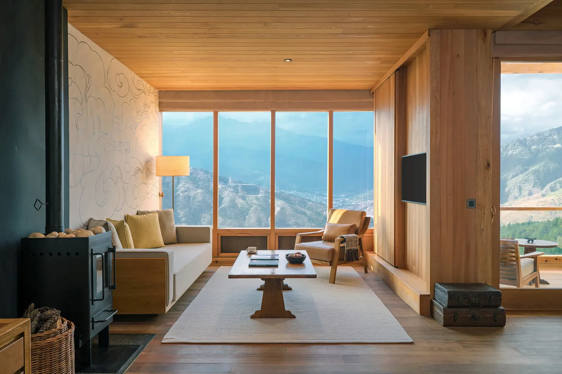 Living room with large floor-to-ceiling windows showing mountain views, wooden walls and ceiling, a white sofa with yellow cushions, a wooden coffee table with books and a bowl, a beige armchair with a throw blanket, a tall lamp, and a black wood-burning stove with stones on top.