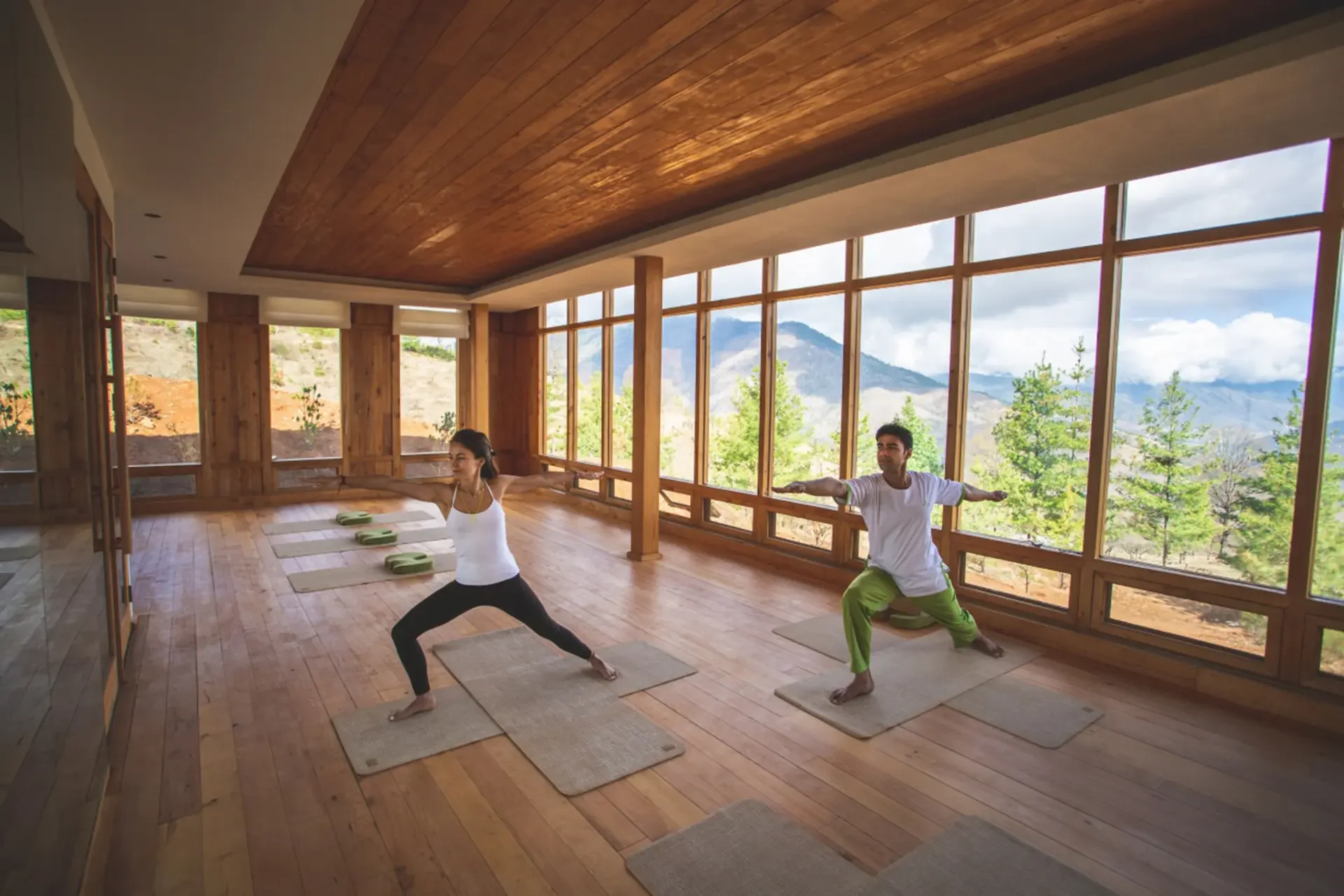 Two people practicing yoga in a spacious room with large windows and a mountain view, with yoga mats on a wooden floor.