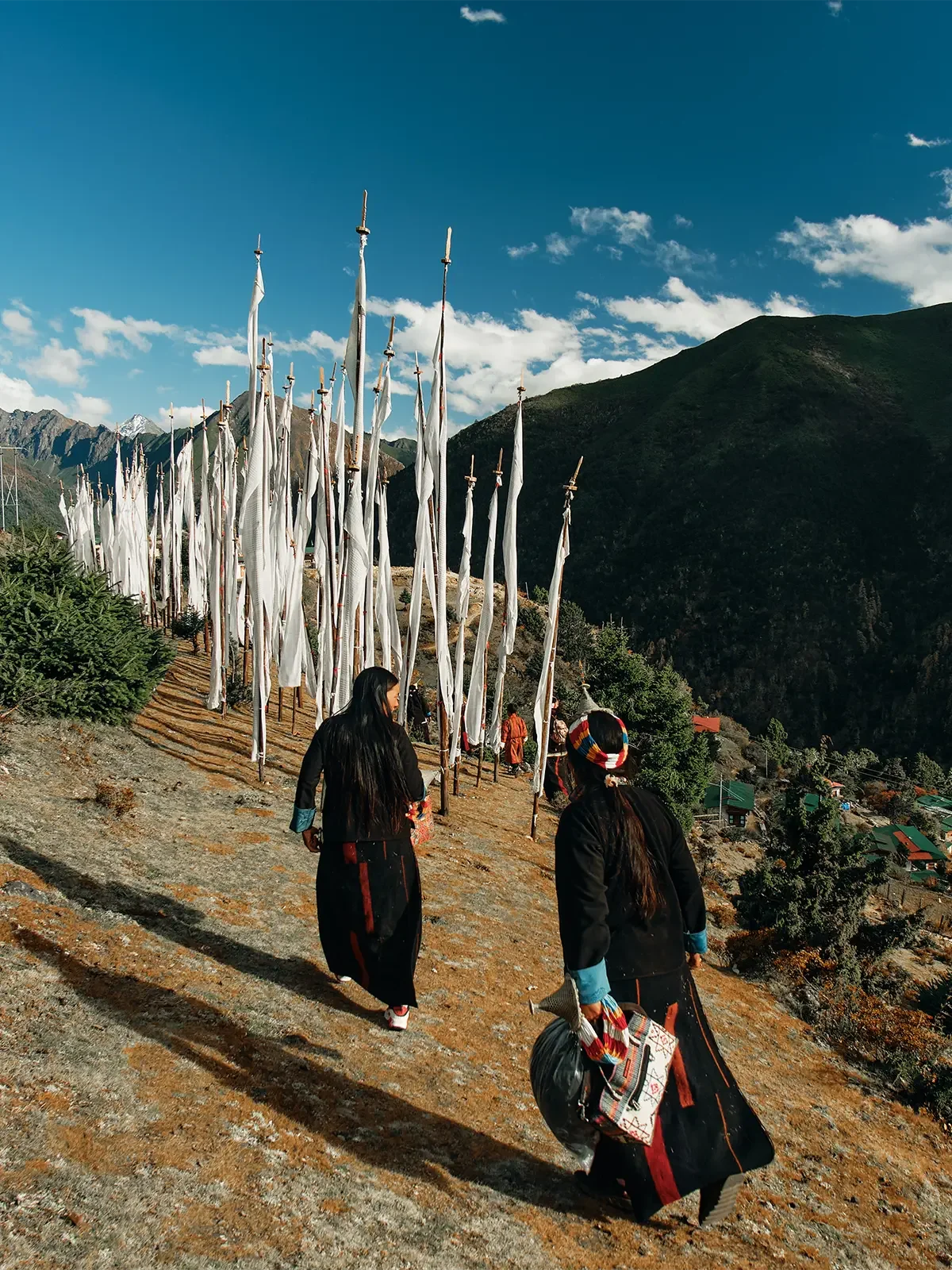 People walking uphill on a trail lined with tall, white prayer flags with mountains in the background and a partly cloudy sky above.