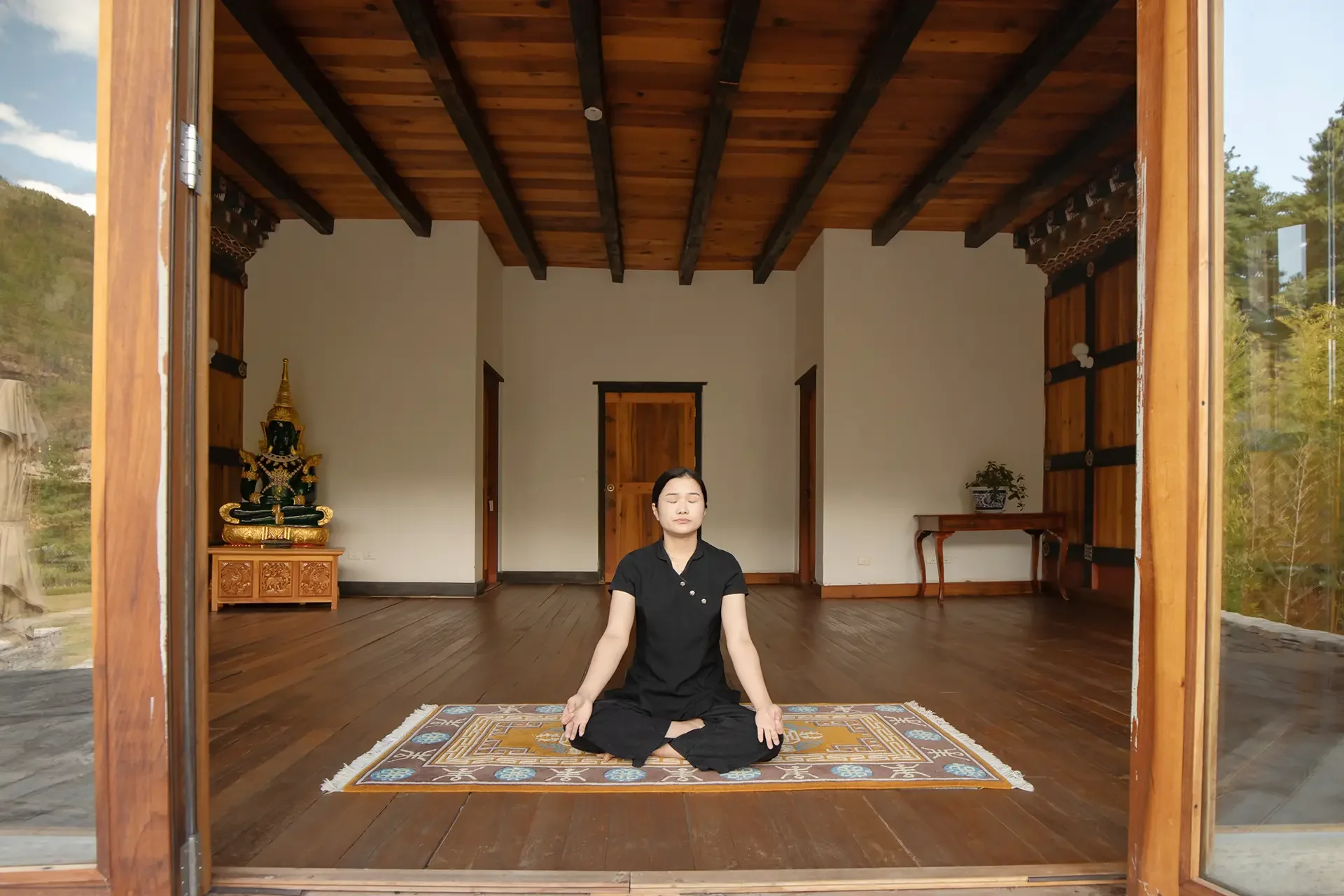 A woman practicing meditation or yoga in a room with wooden floors and a wooden ceiling, sitting cross-legged on an ornate rug with her eyes closed, surrounded by traditional decor including a statue on a small cabinet to the left and a plant on a table to the right