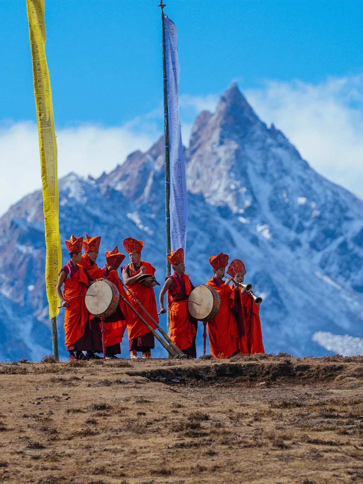Group of monks wearing red robes and traditional hats, playing drums and wind instruments, standing on rocky ground with a snow-capped mountain in the background.