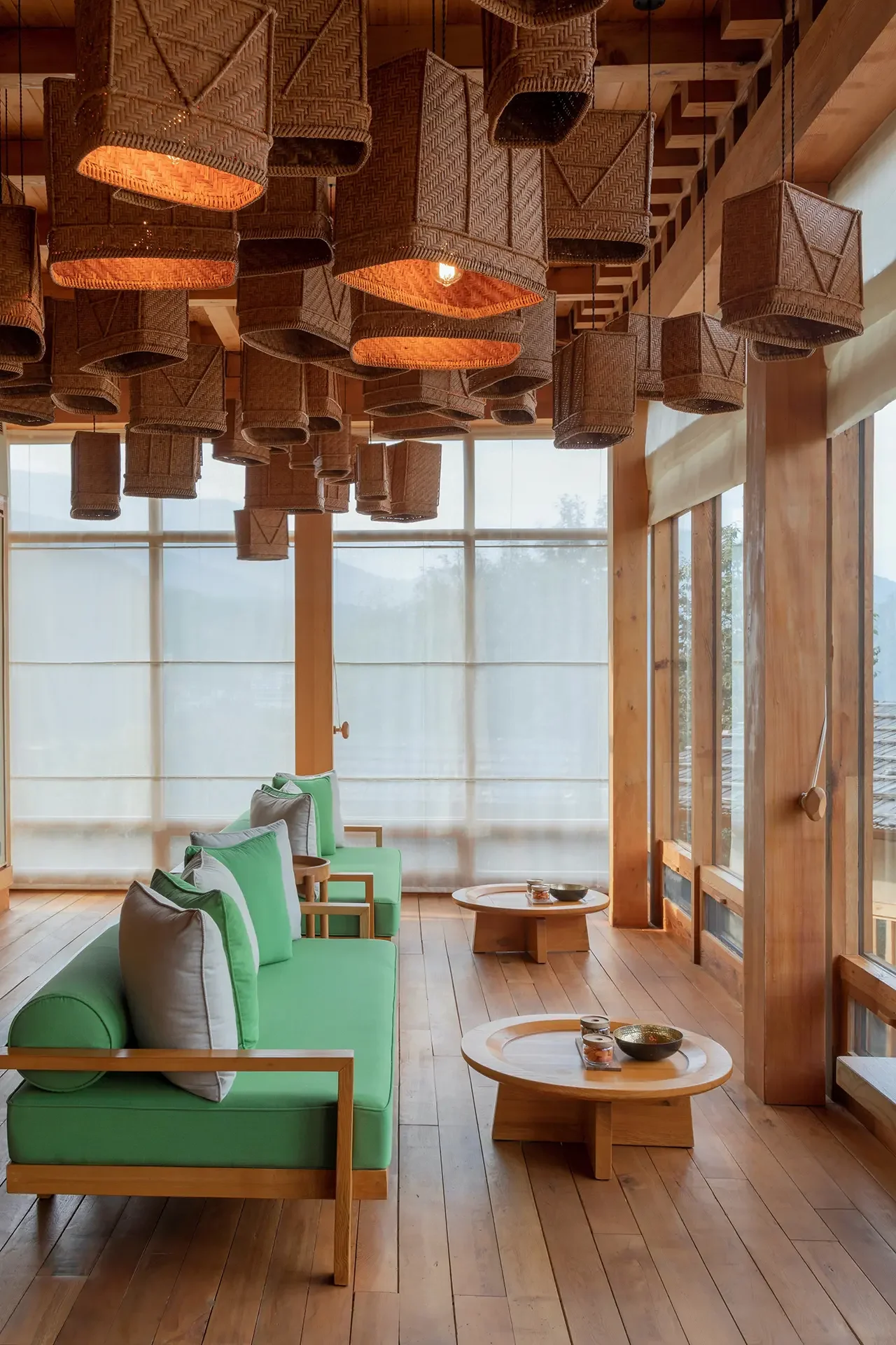 A room with large windows, wooden floor, and wooden beams, decorated with green sofas, cozy cushions, and round wooden coffee tables, eye-catching hanging woven wicker light fixtures, and a scenic mountain view outside.