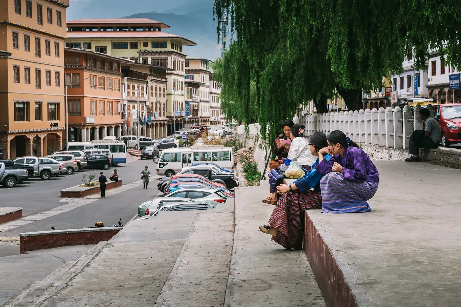 People sitting on a low wall beside a parking lot with cars, trees, and colorful buildings in the background.