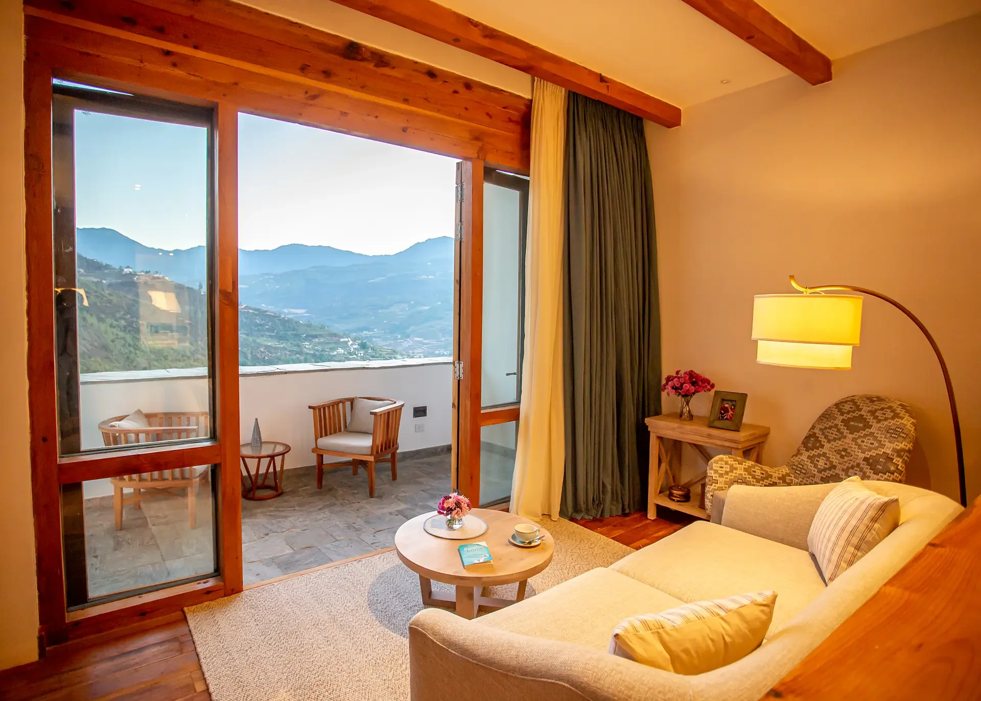 Living room with large glass doors opening to a balcony with mountain views, featuring a beige sofa, a wooden side table with a vase of flowers, a floor lamp, and a small round coffee table with a book and teacup.