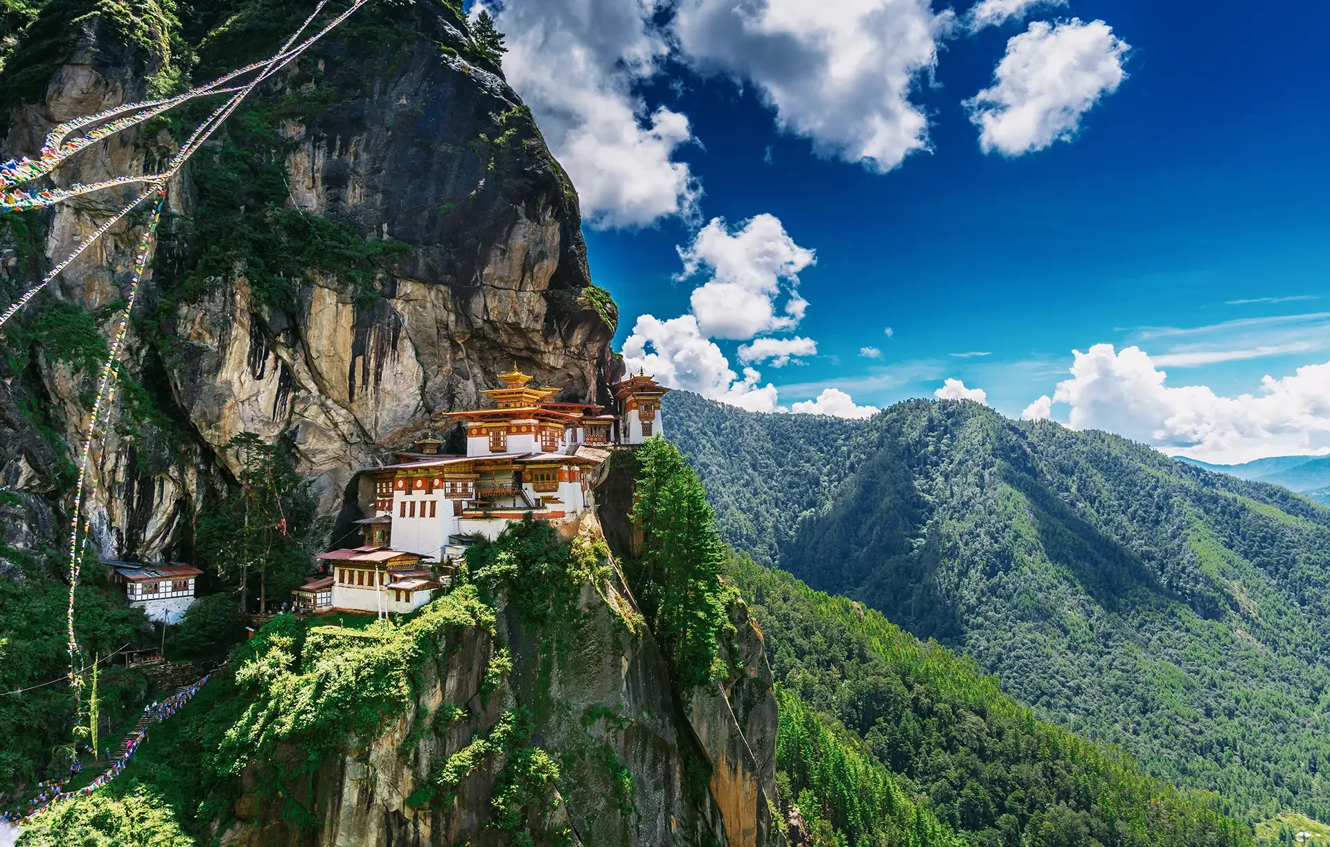 A monastery built into the side of a steep, rocky mountain surrounded by green forest and mountains under a partly cloudy blue sky.
