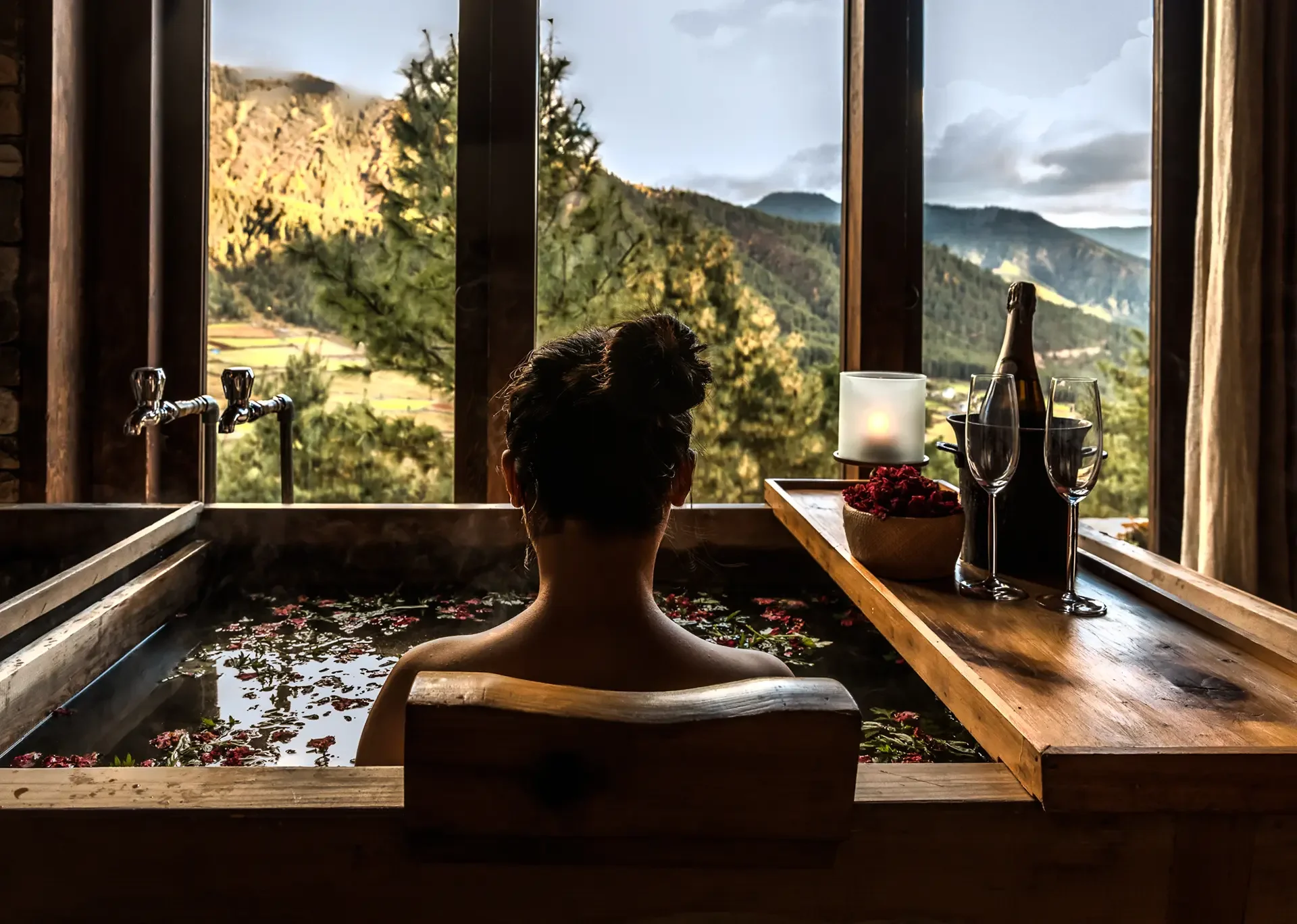 Woman relaxing in a flower-filled bath in a rustic wooden room, overlooking mountains and trees through large windows.