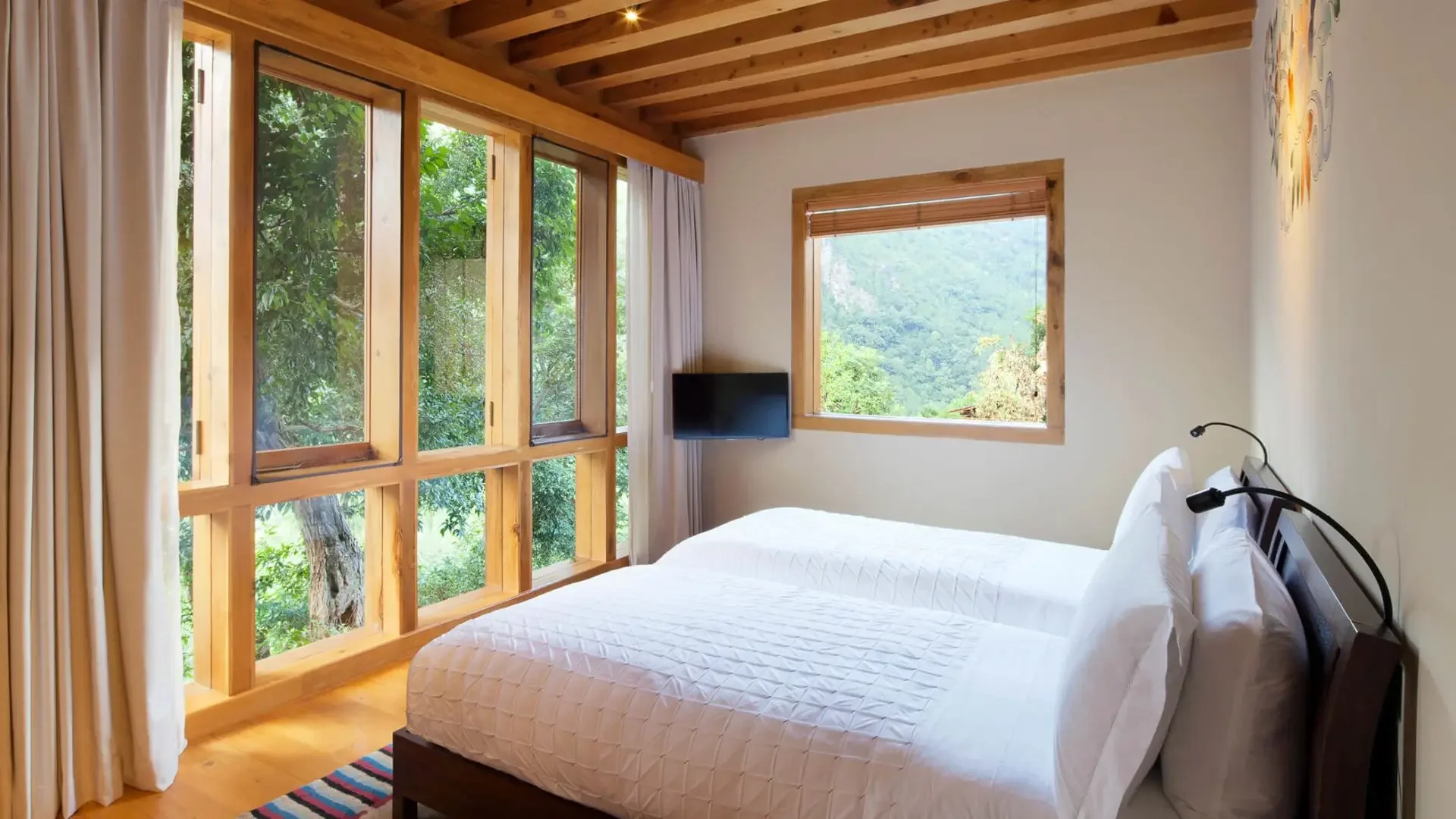 A cozy bedroom with two beds, a large window showing greenery outside, and a smaller window with a scenic mountain view, decorated with wooden accents.