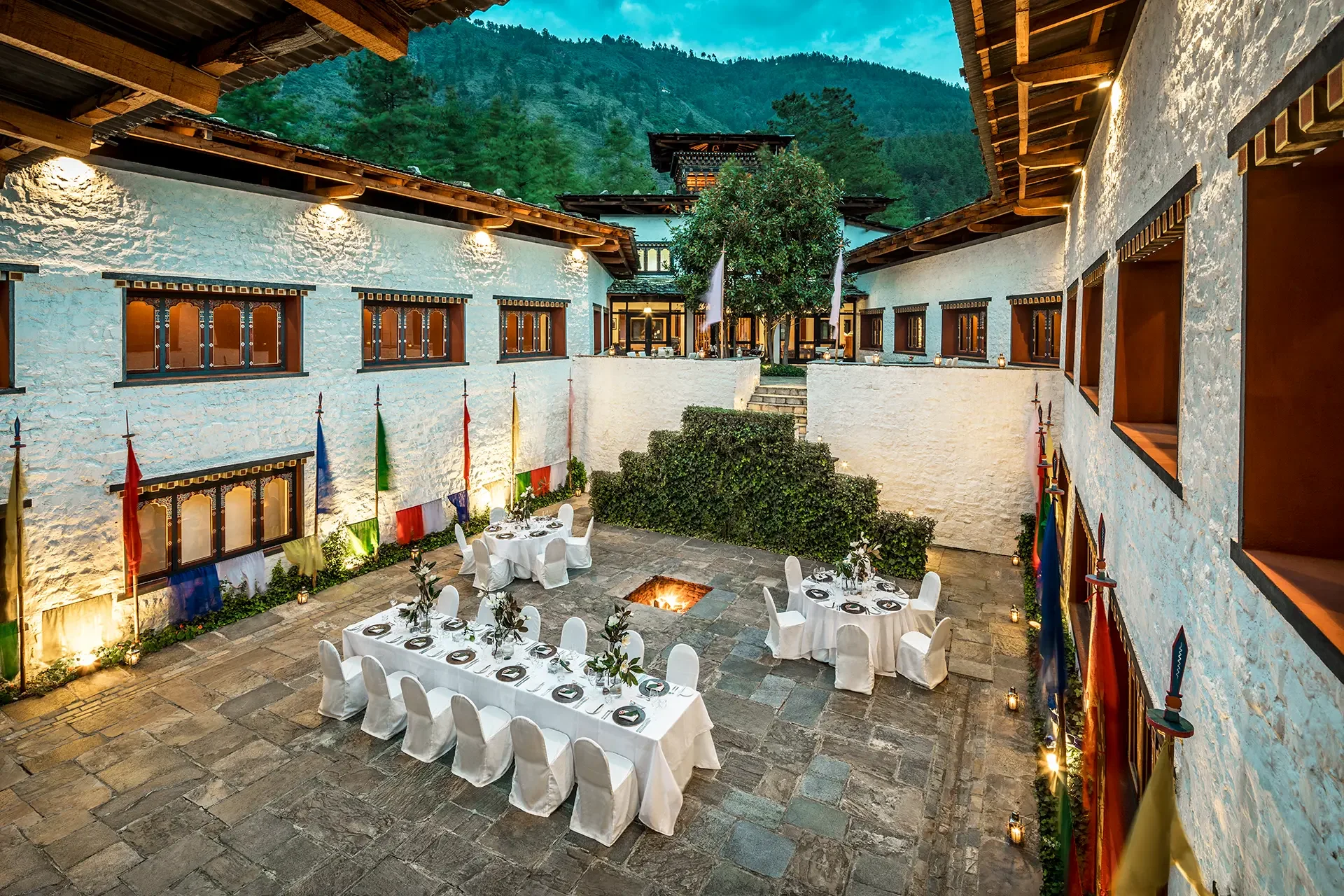 Outdoor courtyard with tables set for a dinner party, surrounded by white stone walls and mountain scenery in the background, decorated with flags and lanterns.