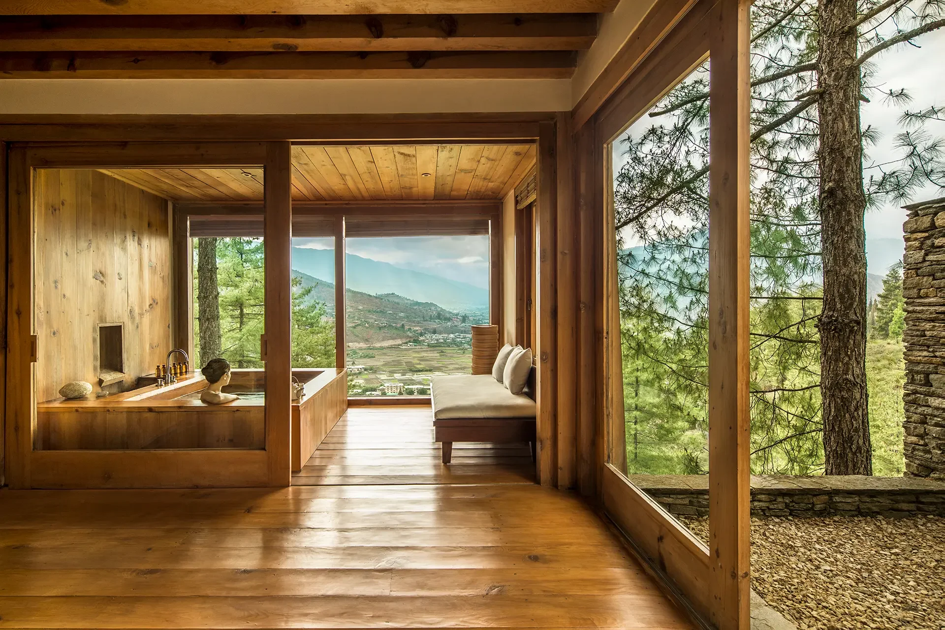 Interior of a wooden cabin with large glass windows overlooking a mountainous landscape with trees and cloudy sky. There is a woman in a bathtub and a cushioned bench inside.