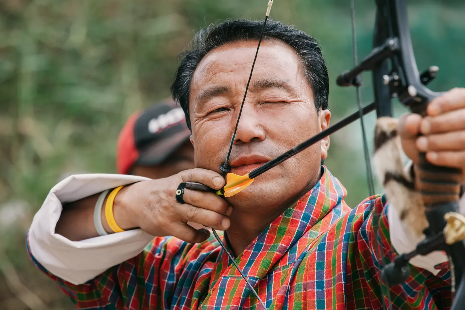 A man in a plaid shirt is aiming a bow and arrow, with another person behind him helping to draw the bowstring.