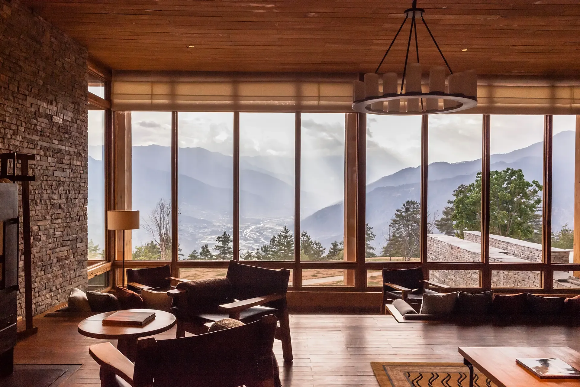 A living room with large floor-to-ceiling windows overlooking mountains and trees, wooden ceiling, stone accent wall, chandelier, and comfortable chairs.