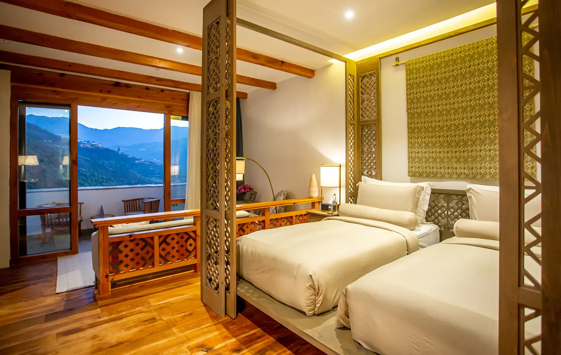 A cozy bedroom with two large beds, a sitting area, and large windows showing a mountain view, decorated with warm lighting and wooden accents.
