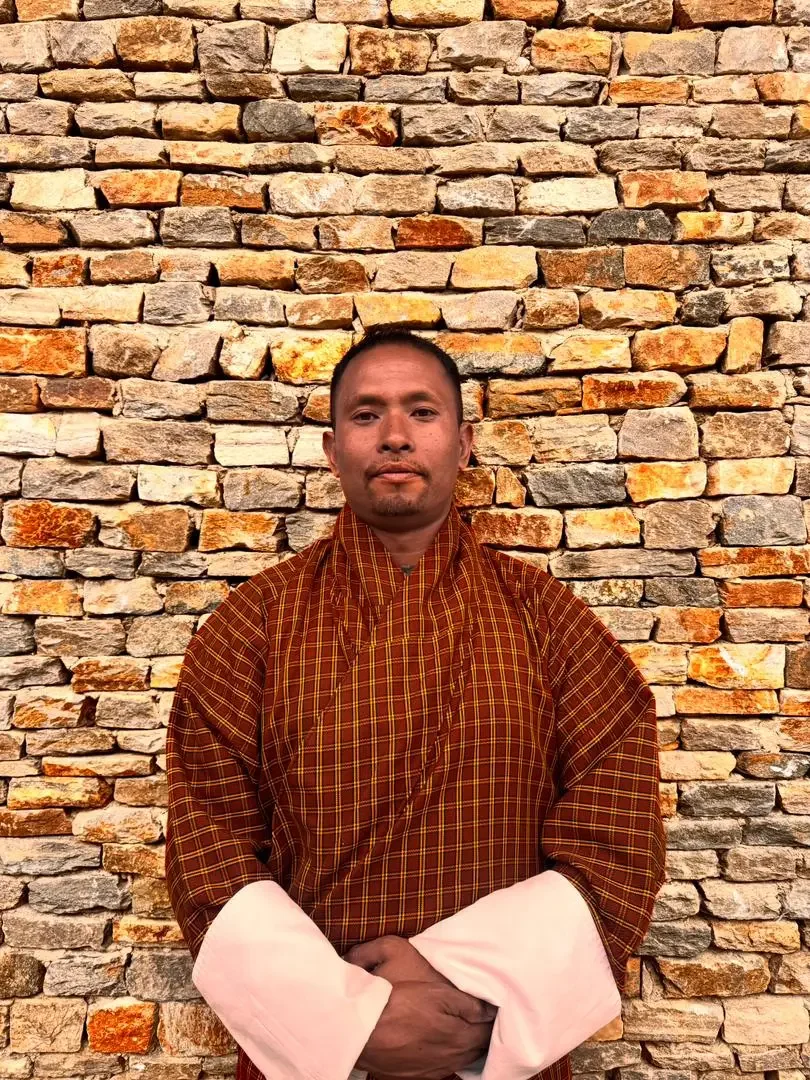 A man dressed in traditional monk or spiritual attire standing in front of a textured stone wall.
