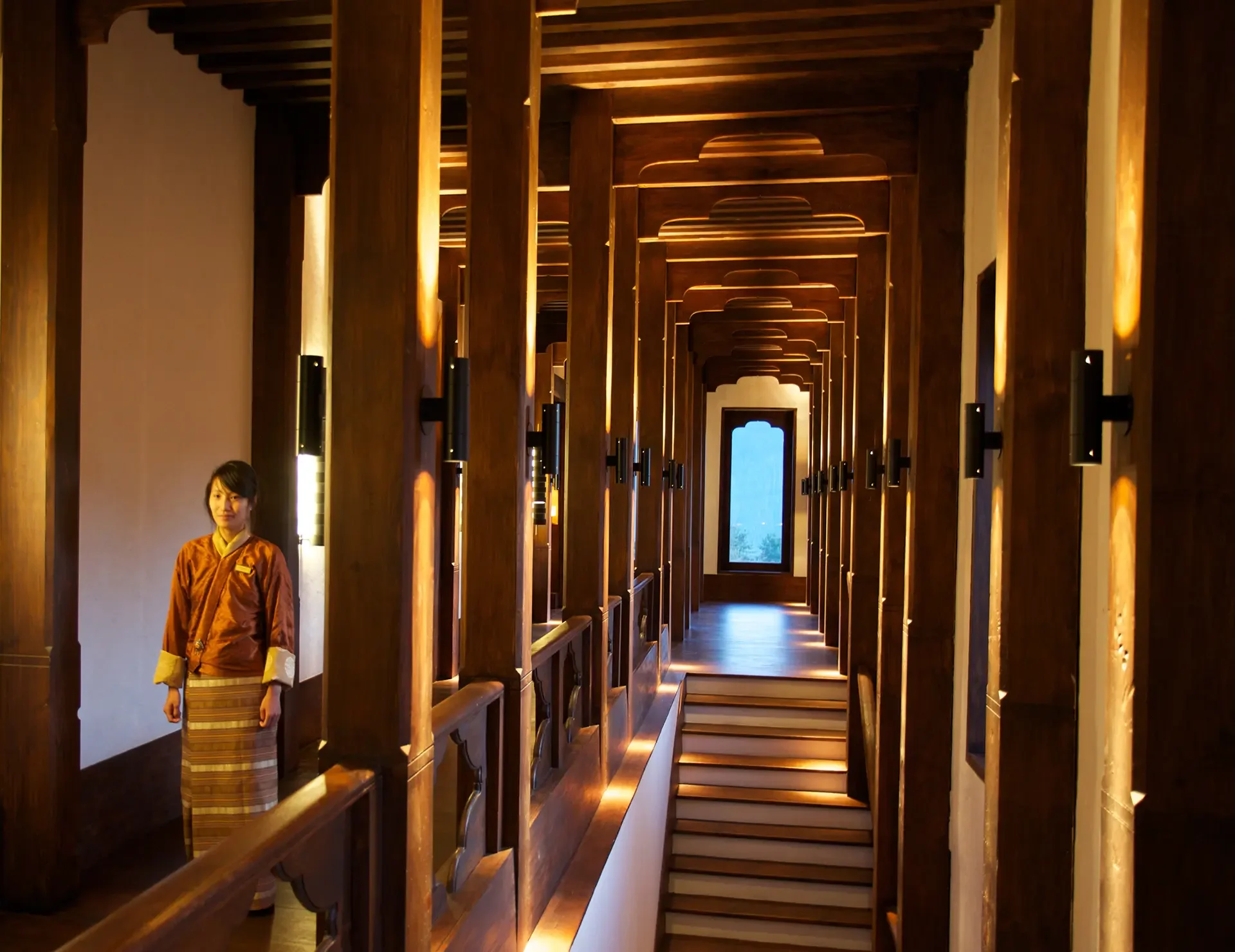 A woman dressed in traditional Asian clothing stands inside a wooden corridor with decorative archways and soft lighting, during daylight.