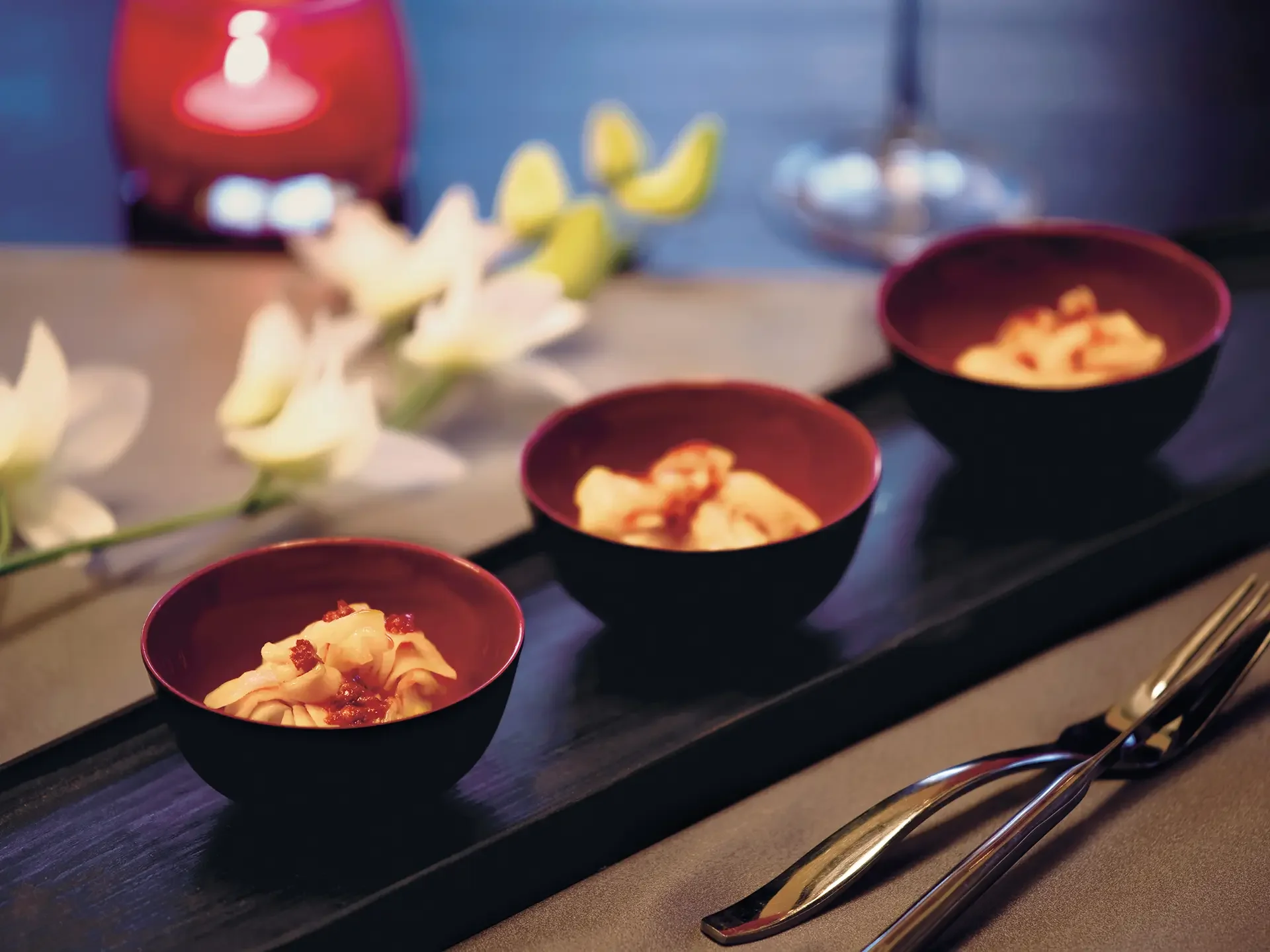 Small black bowls filled with pasta, garnished with red sauce and herbs, placed in a line on a black plate with serving utensils beside them. In the background, there is a candle, some yellow and white flowers, and a glass of red wine, all slightly out of focus.
