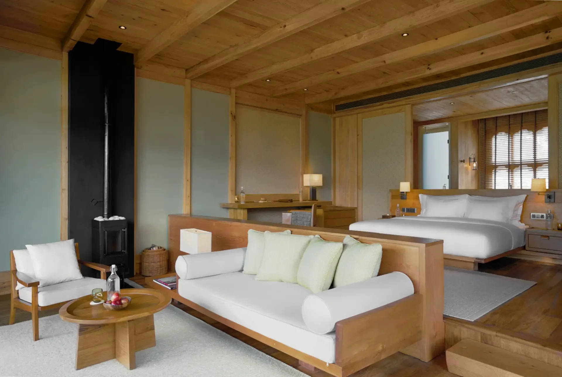 A cozy hotel suite with wooden ceiling and walls, featuring a large bed with white linens, a sitting area with white armchairs and a wooden coffee table with fruit, a small desk with a water bottle, and warm lighting.