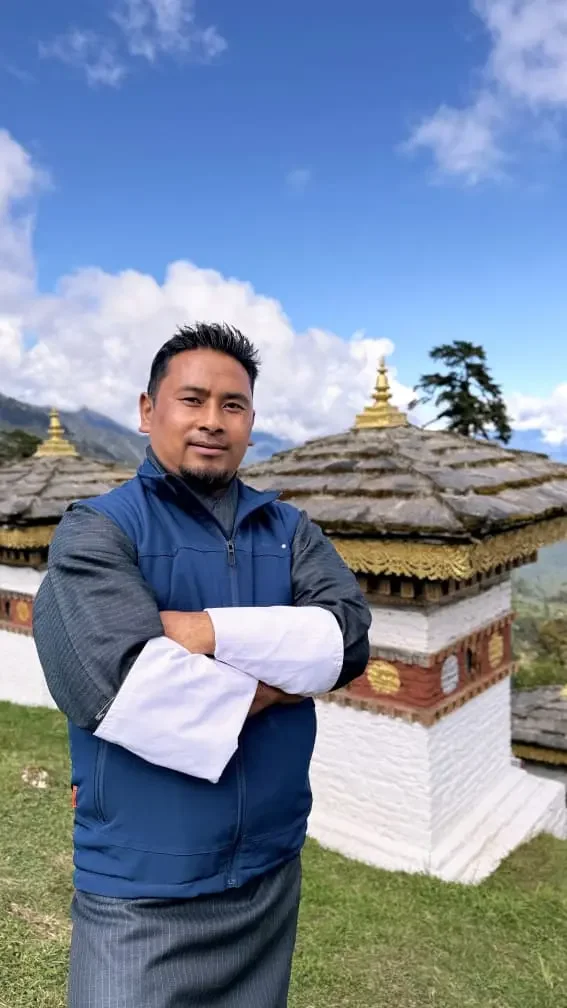 A man standing outdoors with crossed arms, wearing a blue and gray jacket, in front of traditional Asian-style buildings with layered roofs and golden decorative elements, under a partly cloudy sky.