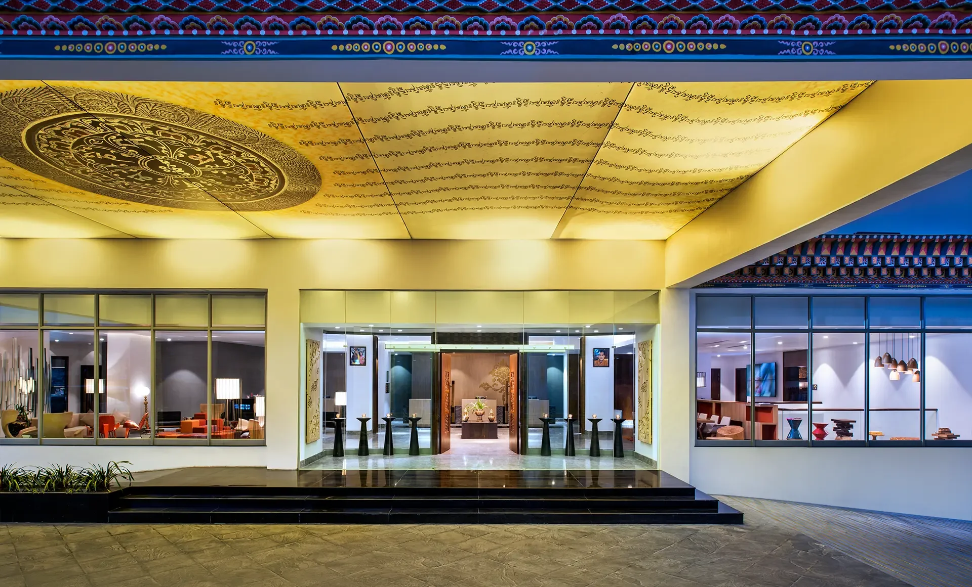 Hotel lobby with large illuminated yellow ceiling panel decorated with ornate gold patterns, a modern entrance with glass windows showing a living room with chairs and lamps, and a reception desk in the background.