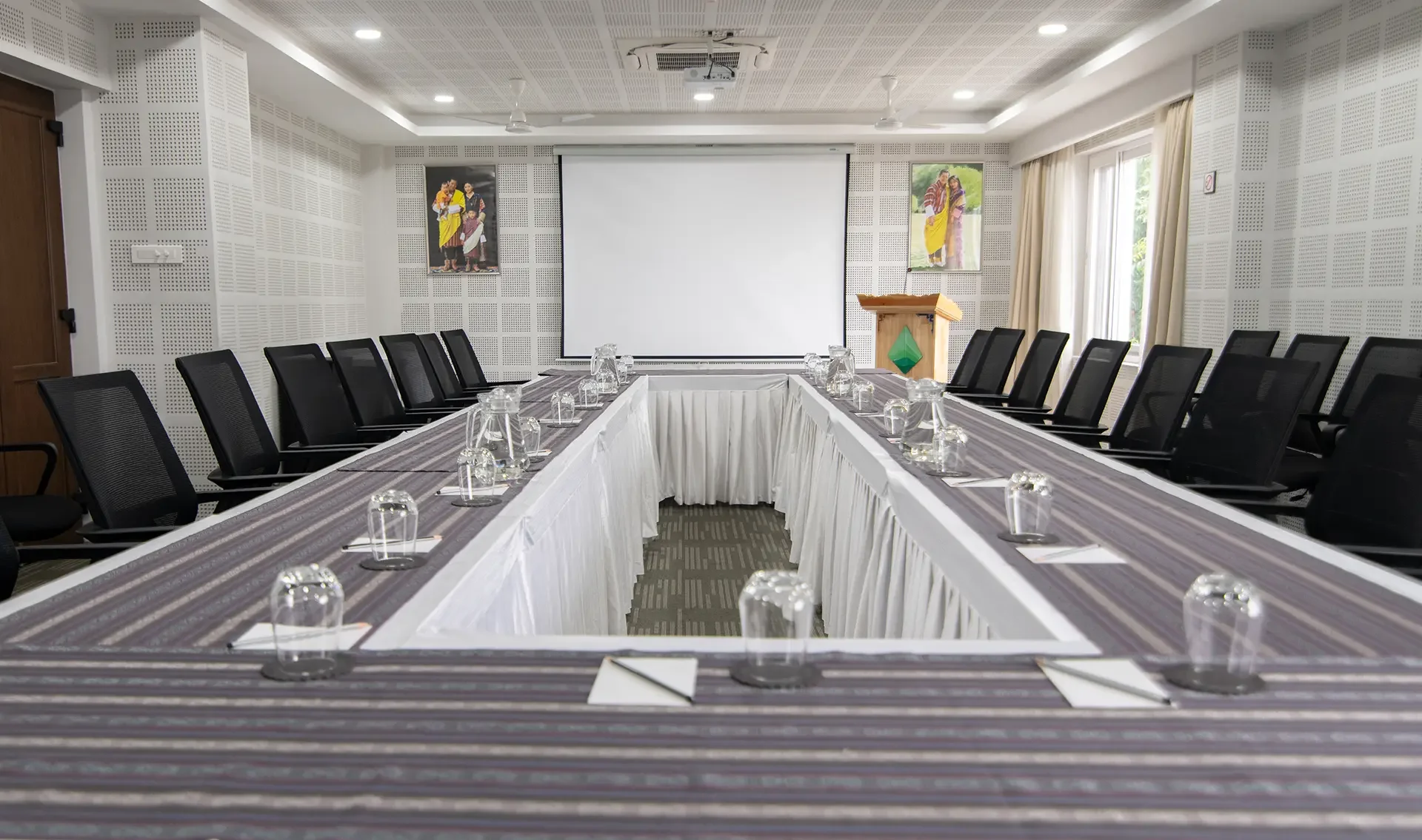 Conference room with a U-shaped table setup, black chairs, glasses, notepads, and a projection screen at the front. Portraits of Indian people hang on the walls, and large windows with curtains let in natural light.