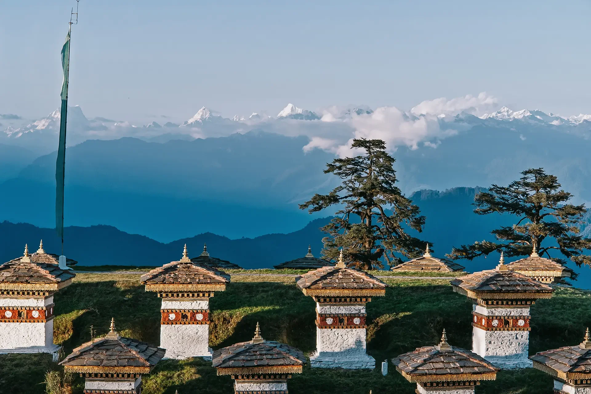 Landscape view of snow-capped mountains in the distance under a clear blue sky. Traditional white temples with ornate, layered roofs are in the foreground, surrounded by trees and greenery.