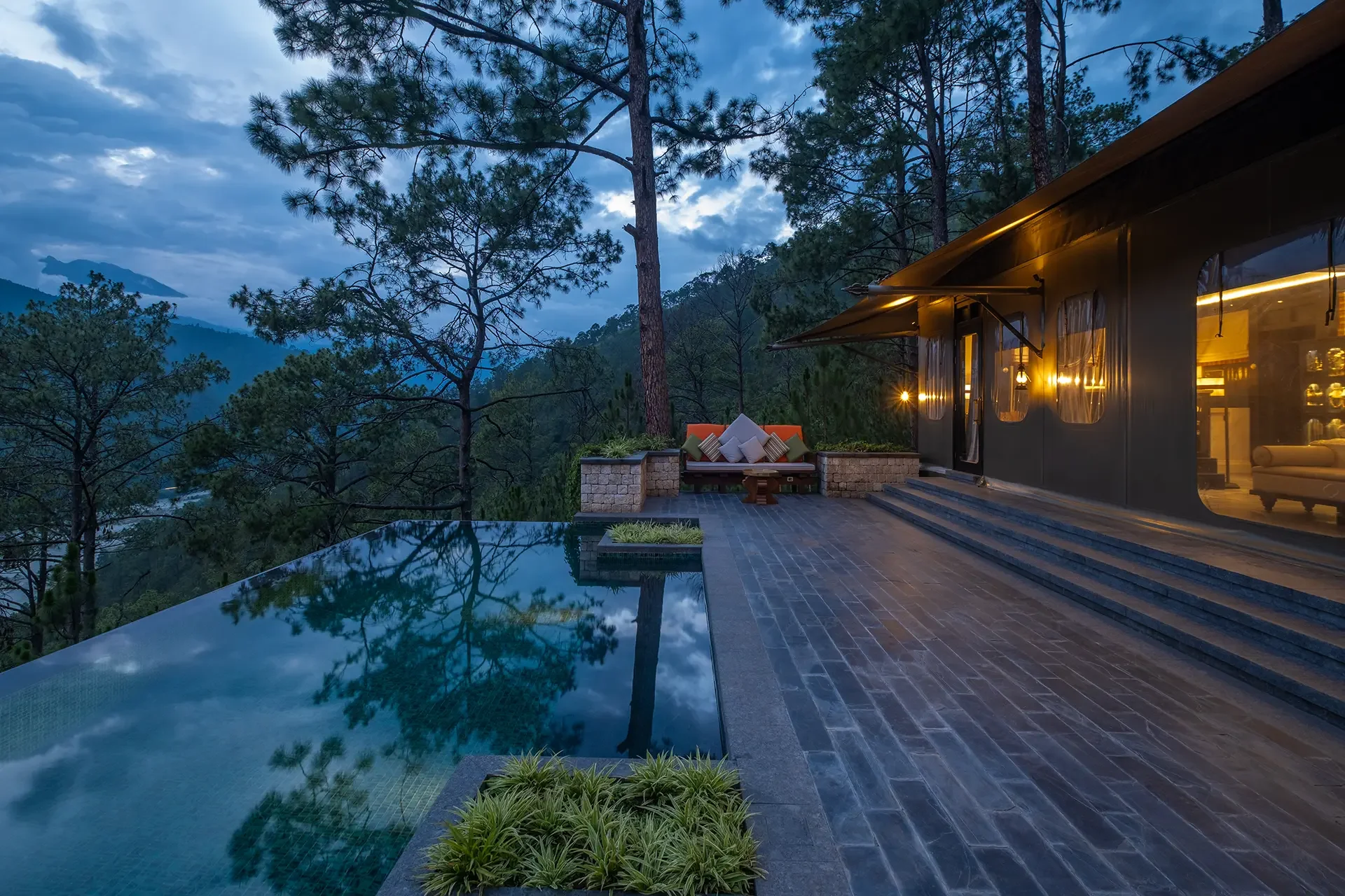 A mountain landscape viewed from a house deck at dusk, featuring trees, a mountain in the background, and an outdoor seating area with cushions. The house has large windows and warm lighting inside. There is a reflecting pool in the foreground.