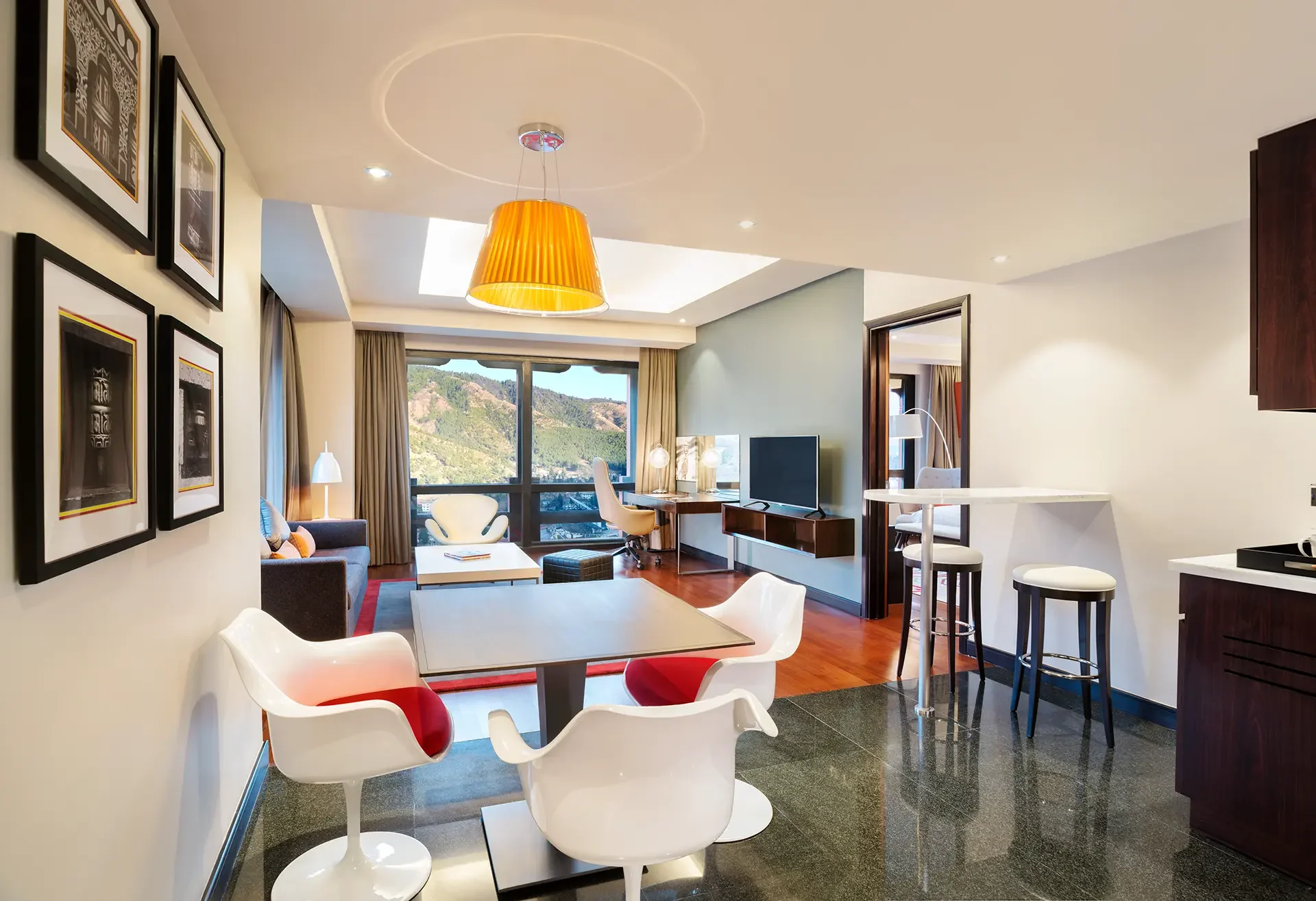 Modern living room with large window showing mountains, featuring a dining table with white chairs, a TV, a desk with chairs, and decorative framed artworks on the wall, illuminated by ceiling and table lighting.