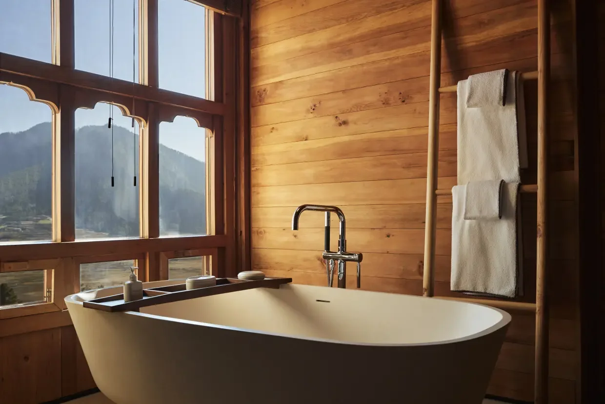 A cozy bathroom with a large bathtub next to a wooden wall and large window showing mountains outside.