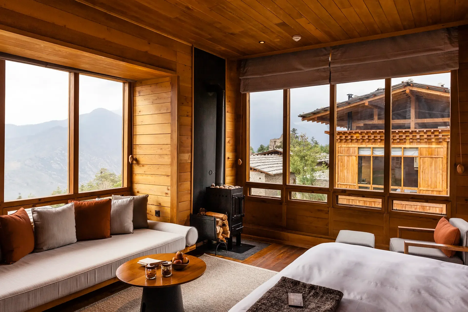 A cozy living room with wooden walls and large windows offering mountain views, includes a white sofa with orange and beige pillows, a small round wooden coffee table with candles, and a wood stove near the corner.